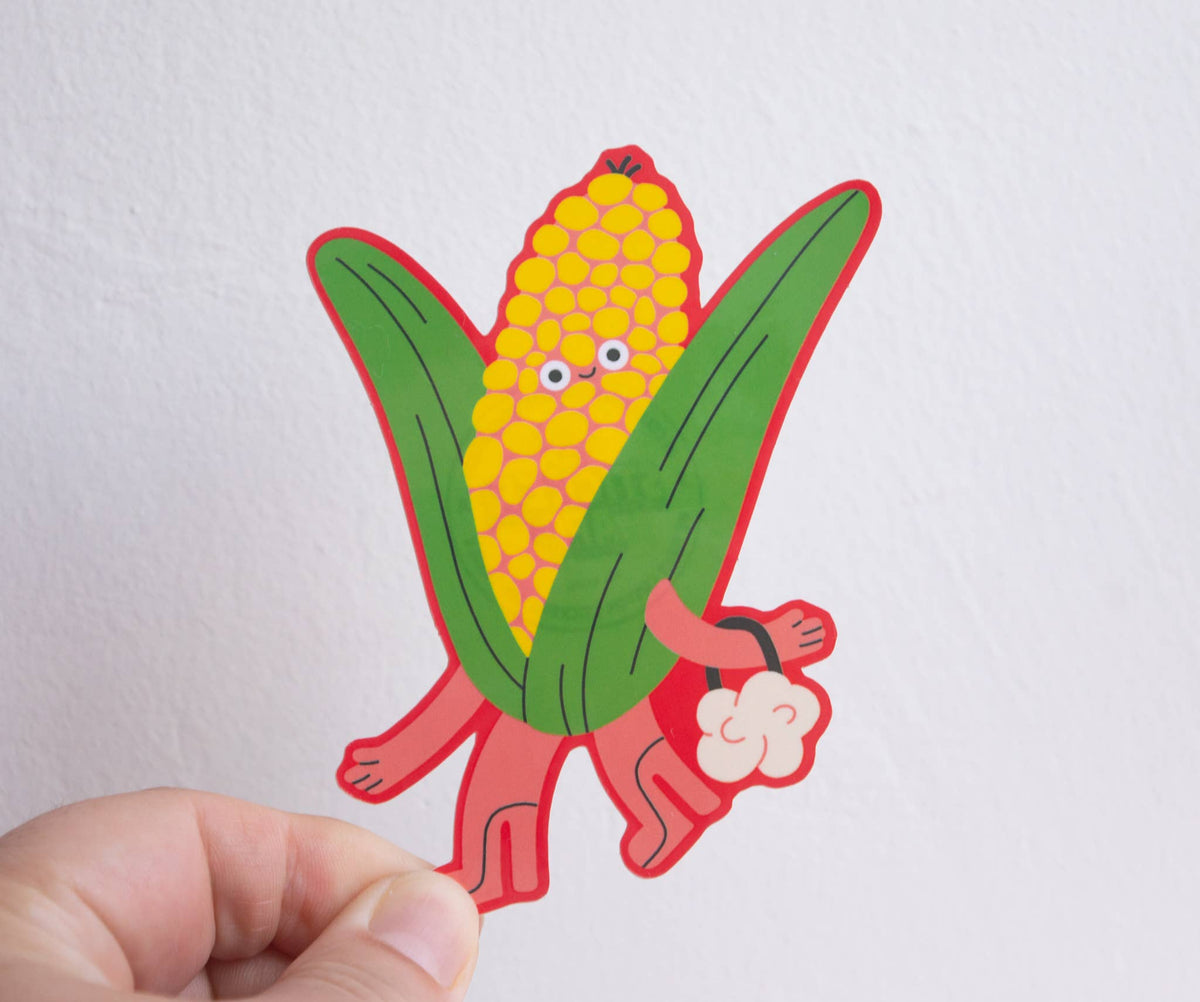 A hand holding a die-cut cartoon sticker of a smiling corn cob with green leaves and red outline, against a plain white wall.