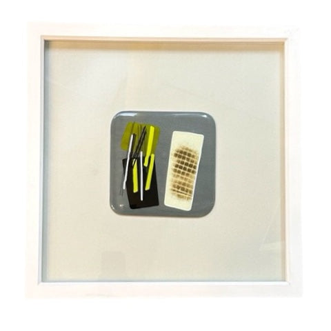 A square fused glass wall hanging, set in a white frame. This glass square is a soft grey, with black and white squares, black and yellow stripes, and crosshatching.