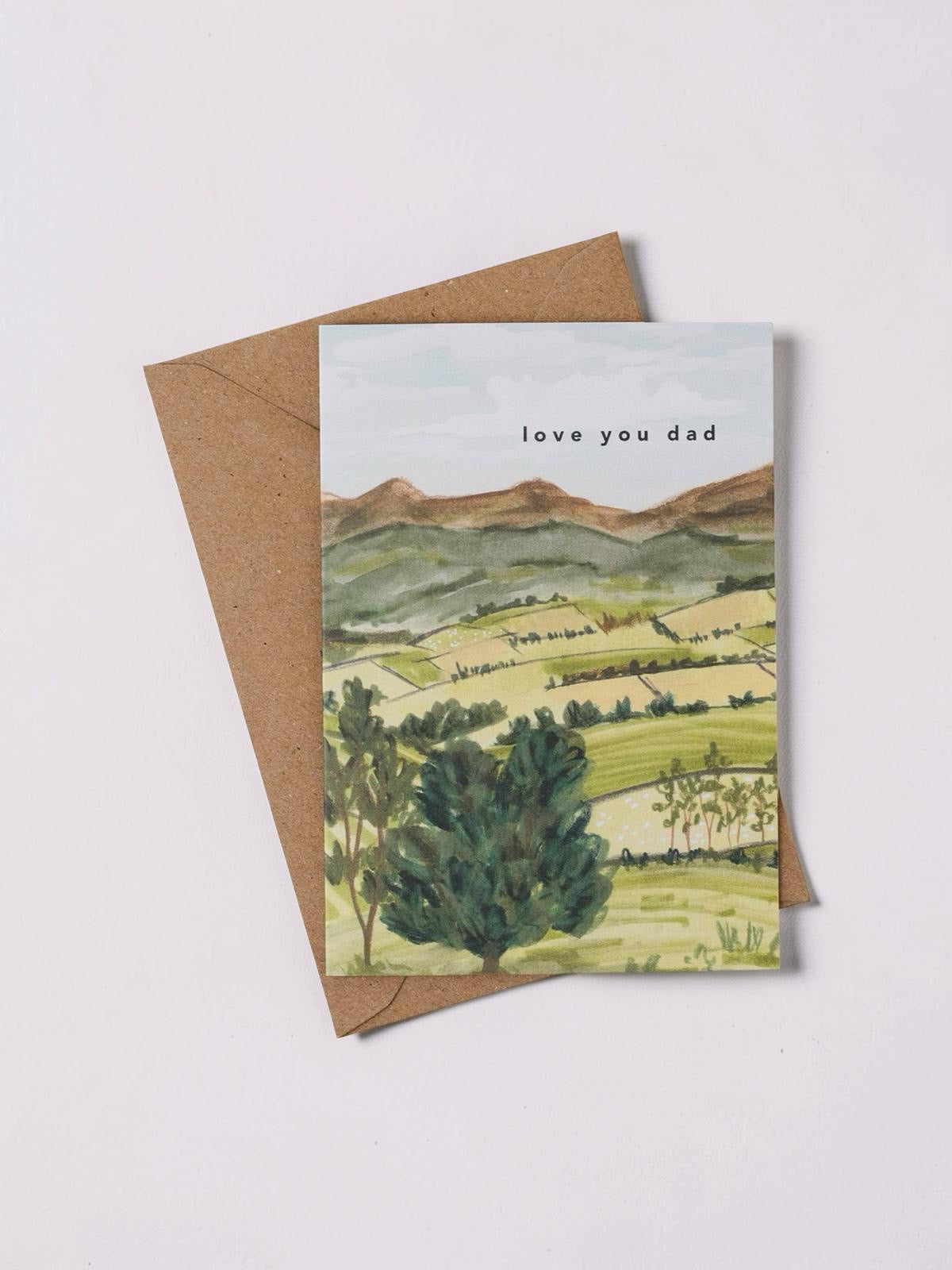 Greeting card with landscape illustration and 'love you dad' text, with a brown envelope.