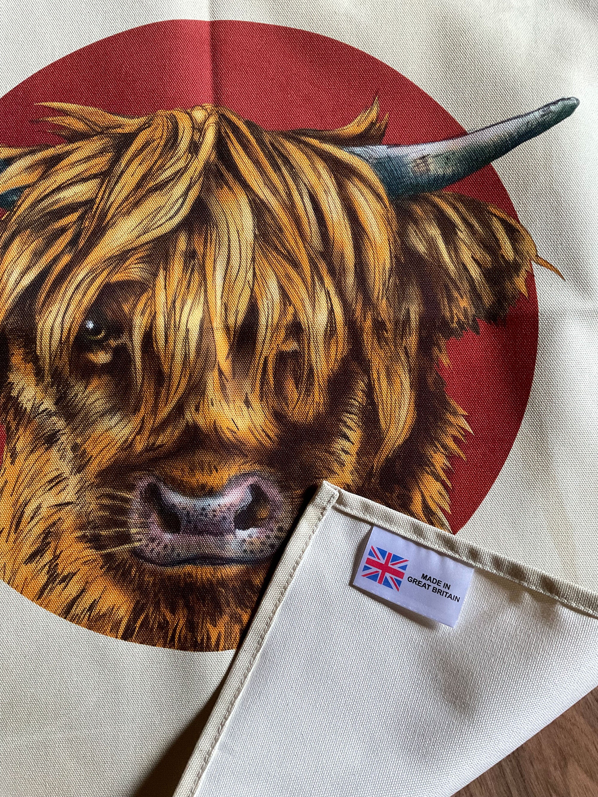 A tea towel featuring an illustration of the face of a highland cow, in a deep red circle against an off-white background. 