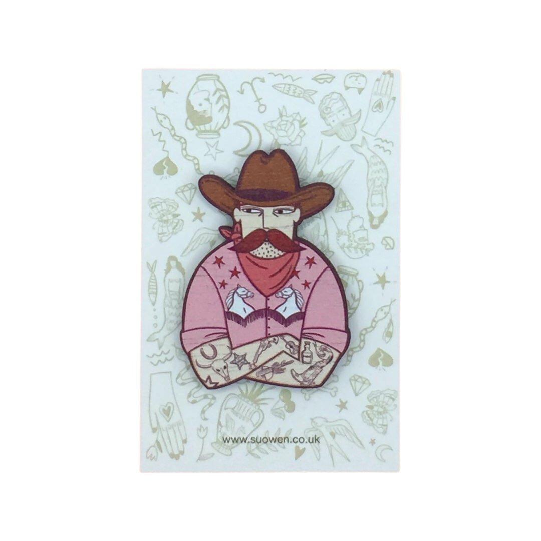 Wooden cowboy brooch featuring a tattooed cowboy wearing a pink shirt with a white horse design, a red neckerchief and a brown cowboy hat.
