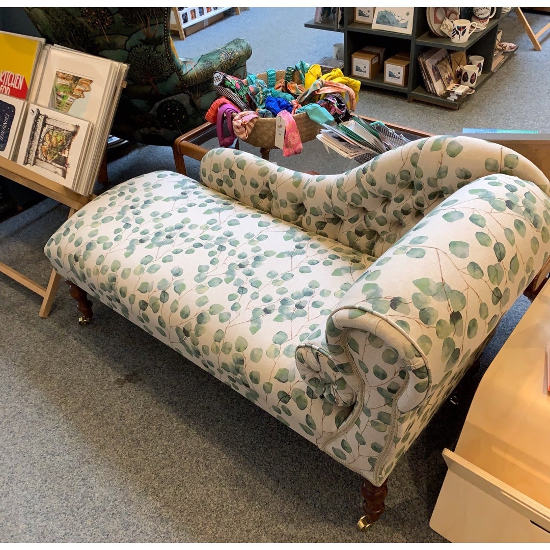 A handcrafted chaise lounge with cream leaves cotton fabric, mahogany stained legs, and brass-colored castors.
