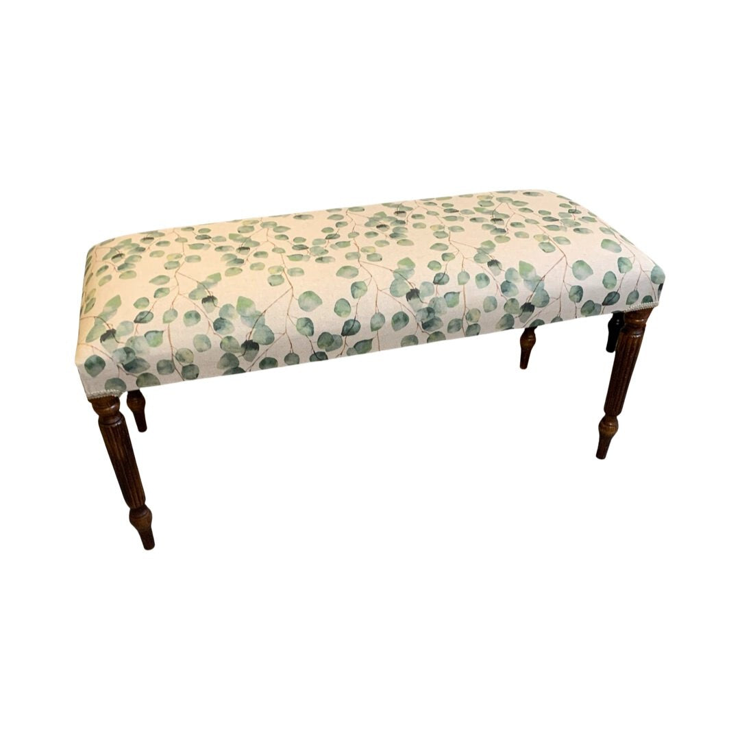 A rectangular bench with mahogany legs and cream linen fabric upholstered in a leaf pattern.