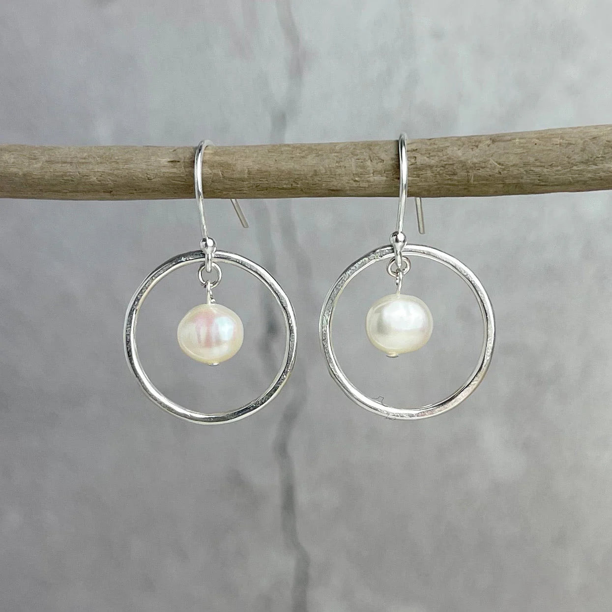 Silver hoop earrings with pearl drops hanging on a branch against a gray background.