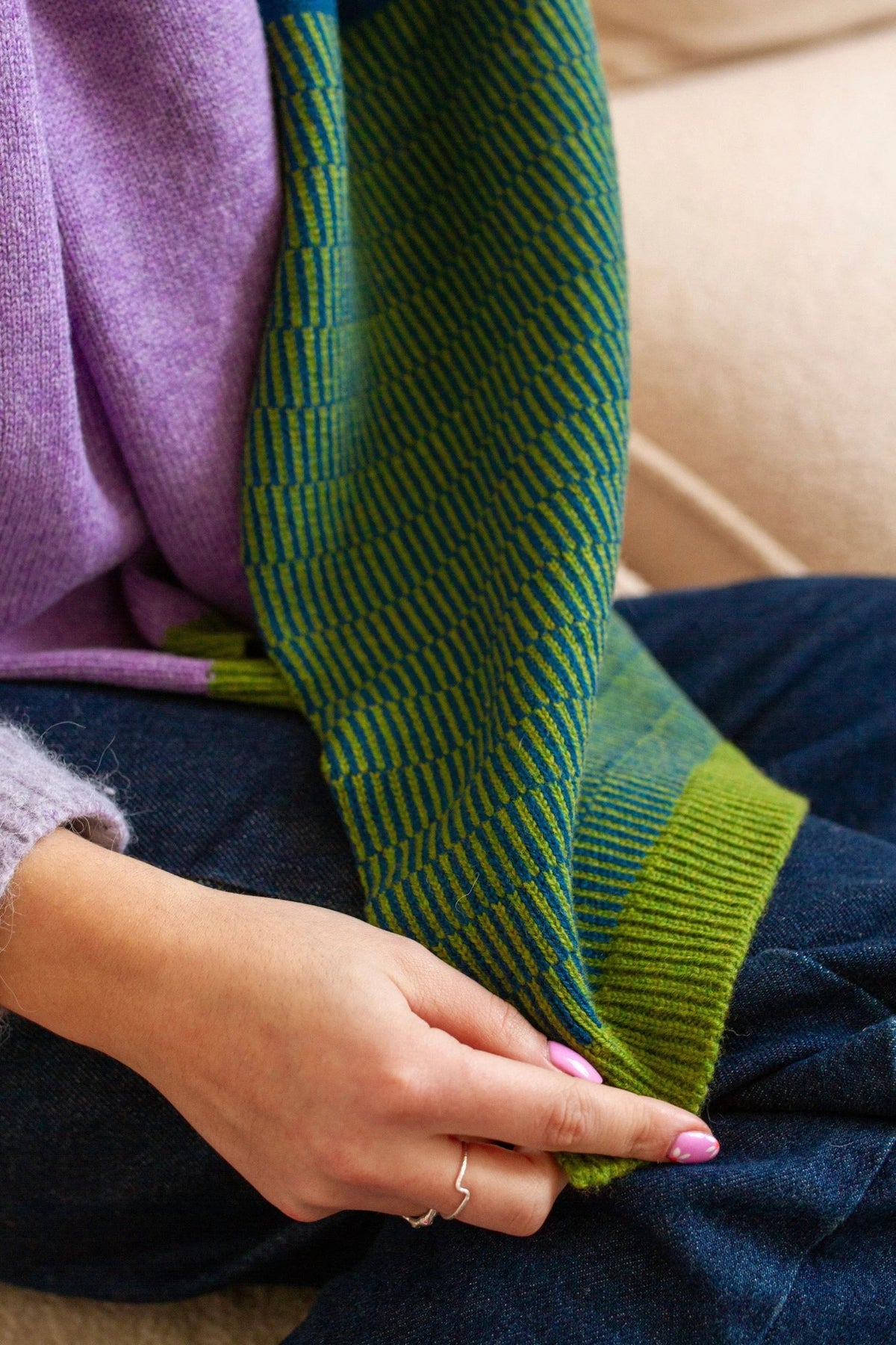 Green knitted scarf being held by a person wearing a purple sweater and blue jeans.