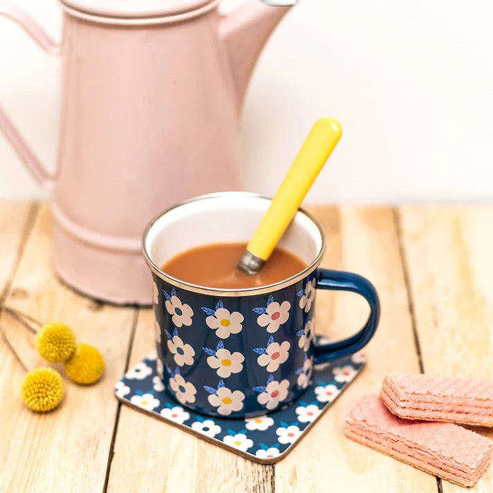 Navy blue mug and coaster with floral pattern on a wooden table with pink teapot, yellow spoon, and cookies.