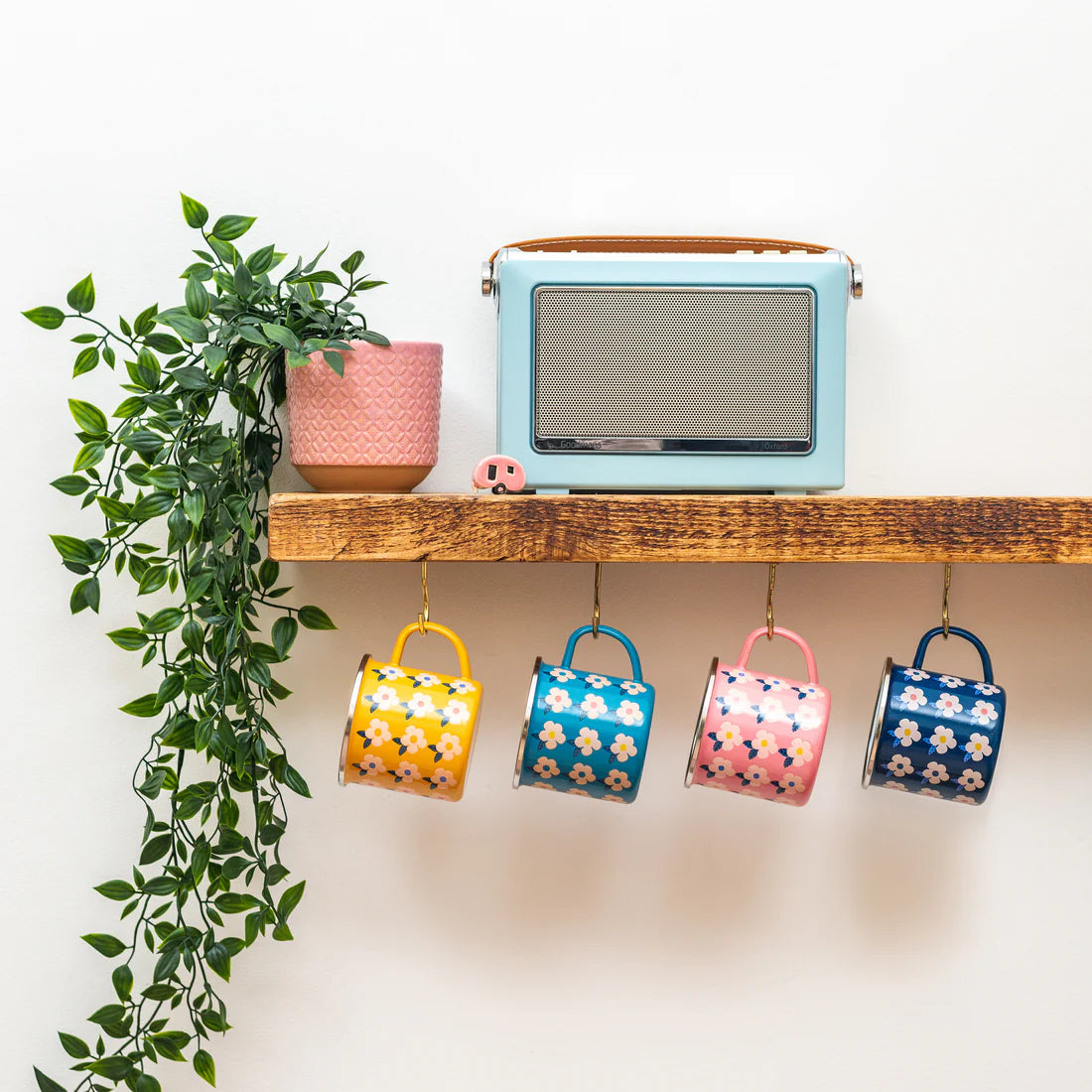 Four colourful patterned ceramic mugs hanging from a wooden shelf, with a small pink plant pot and a light blue radio on the shelf above, against a white wall.