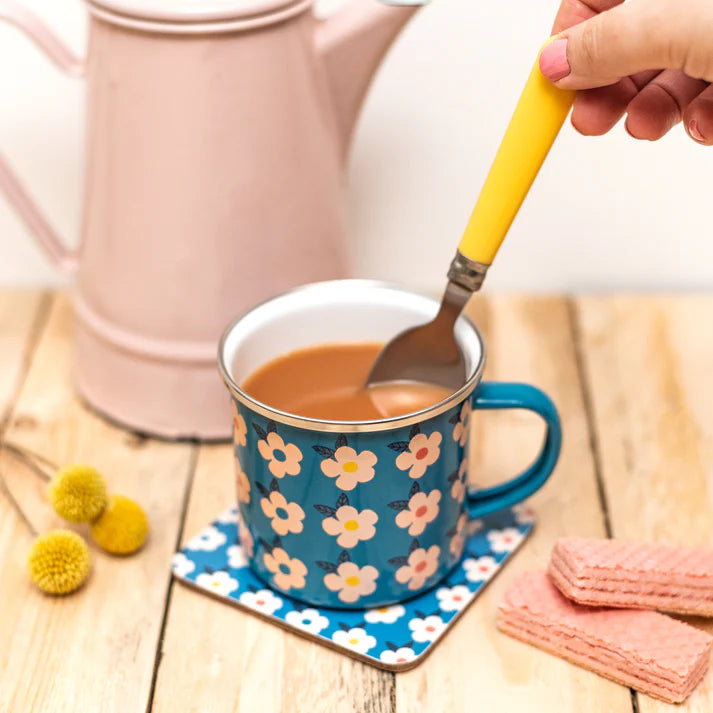 A blue enamel mug with a white and pink floral pattern, placed on a matching coaster. A hand holds a yellow-handled spoon in the mug, with a pink teapot in the background on a wooden table.