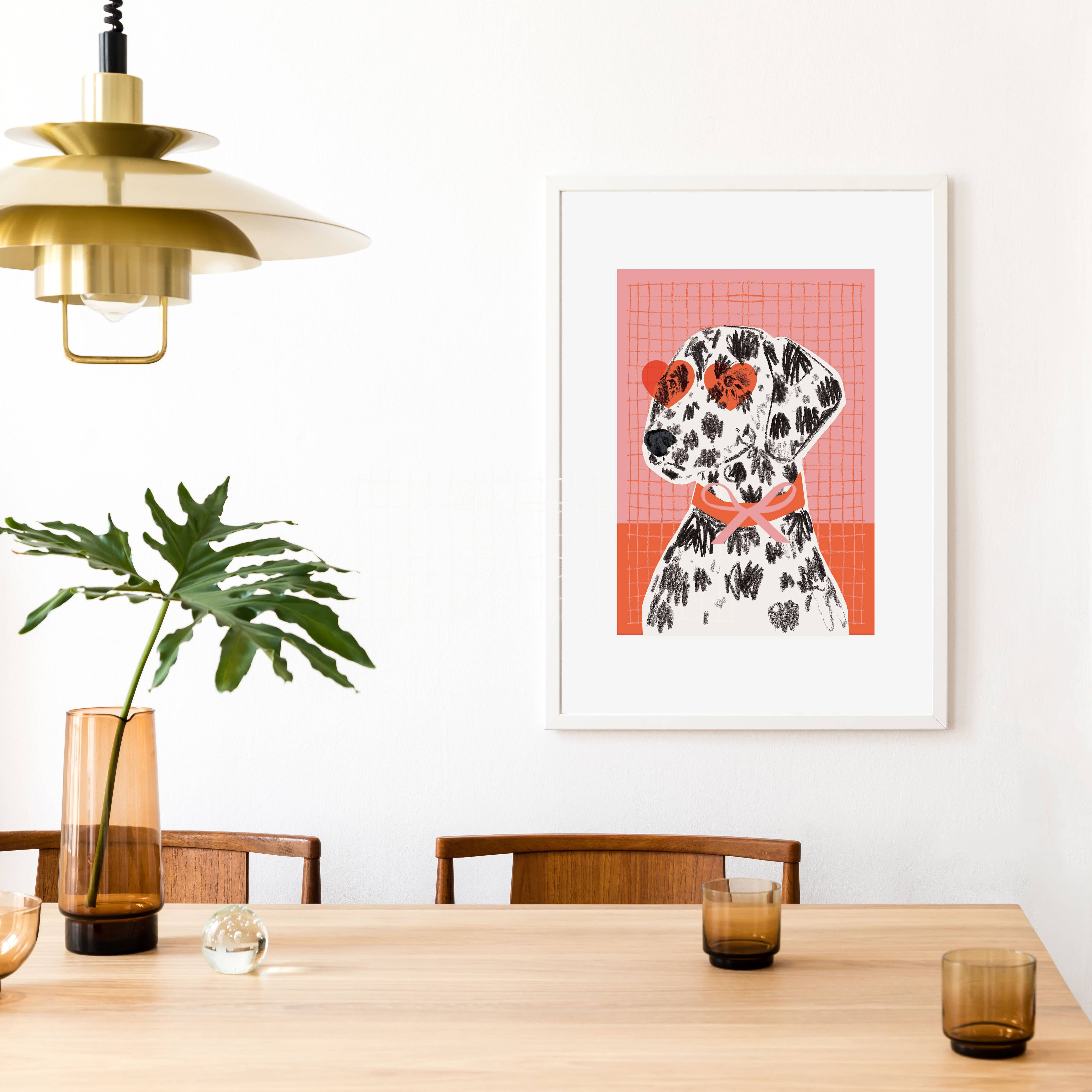A framed art print featuring a Dalmatian dog on a pink and orange grid background, hanging on a white wall in a modern dining room setting with a wooden table, chairs, and a hanging brass lamp.