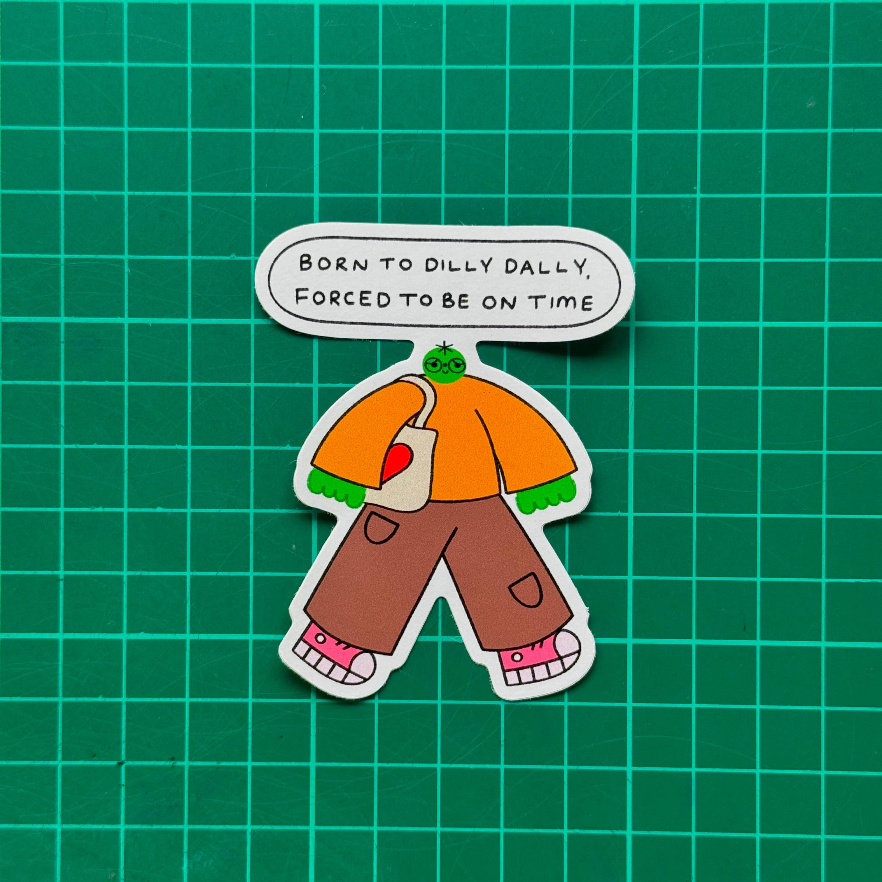 A cartoon-style sticker of a green-skinned character with an orange shirt, brown pants, pink shoes, and a white tote bag, with a speech bubble above reading 'BORN TO DILLY DILLY, FORCED TO BE ON TIME', on a green grid background.