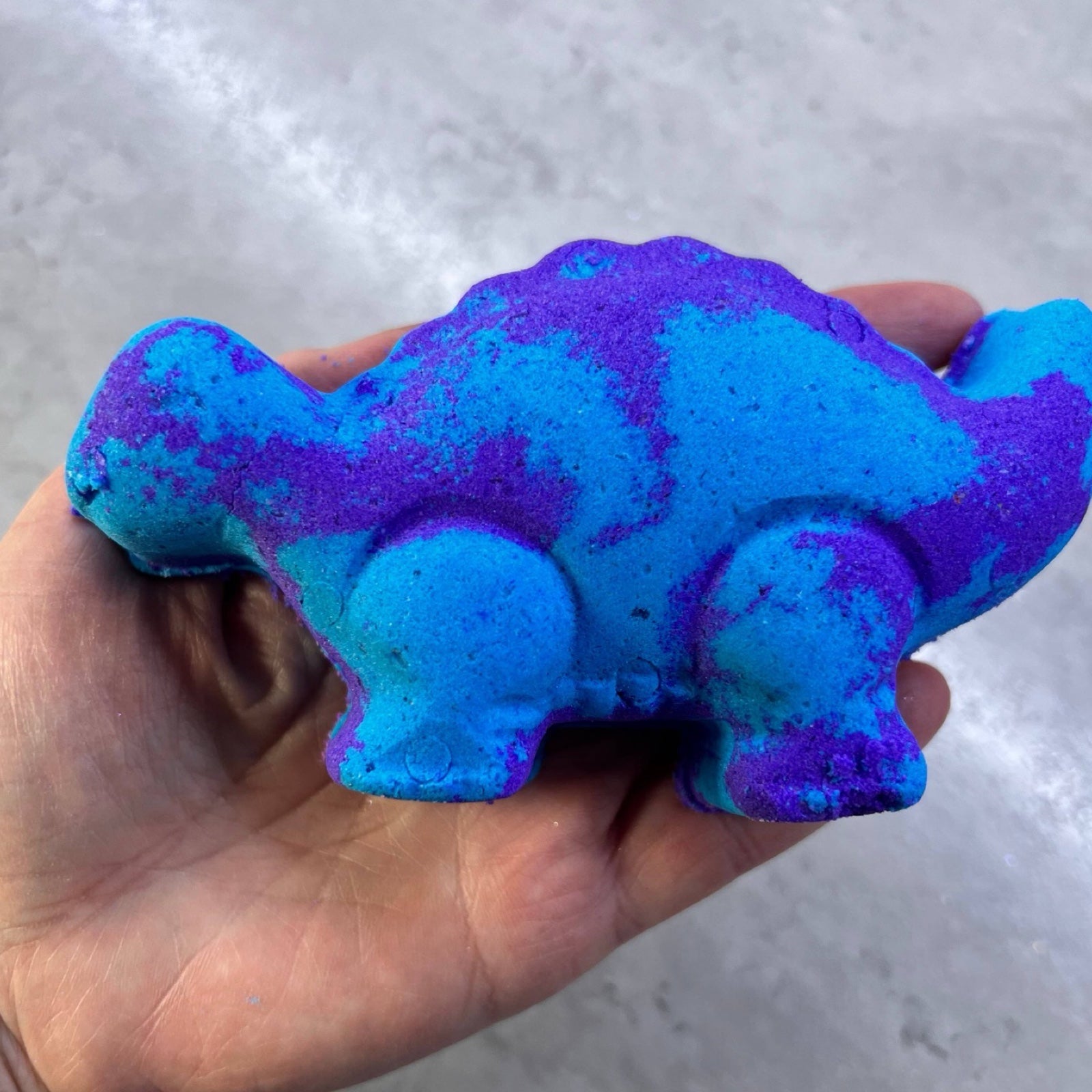 A blue and purple dinosaur-shaped bath bomb held in a ladies hand