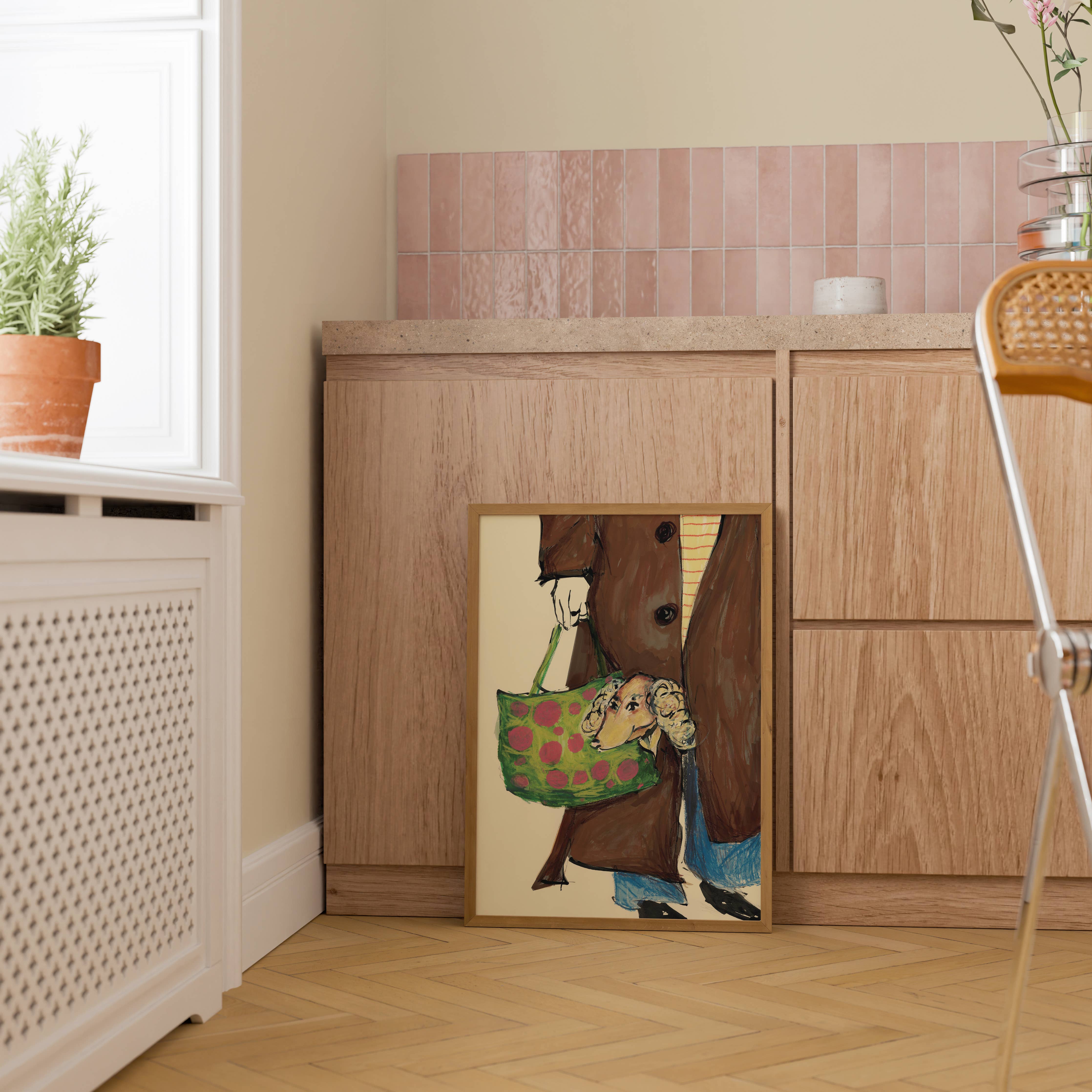 A framed art print leaning against a light wood cabinet in a kitchen setting. The print features a whimsical illustration of a brown bear holding a green and pink patterned bag.
