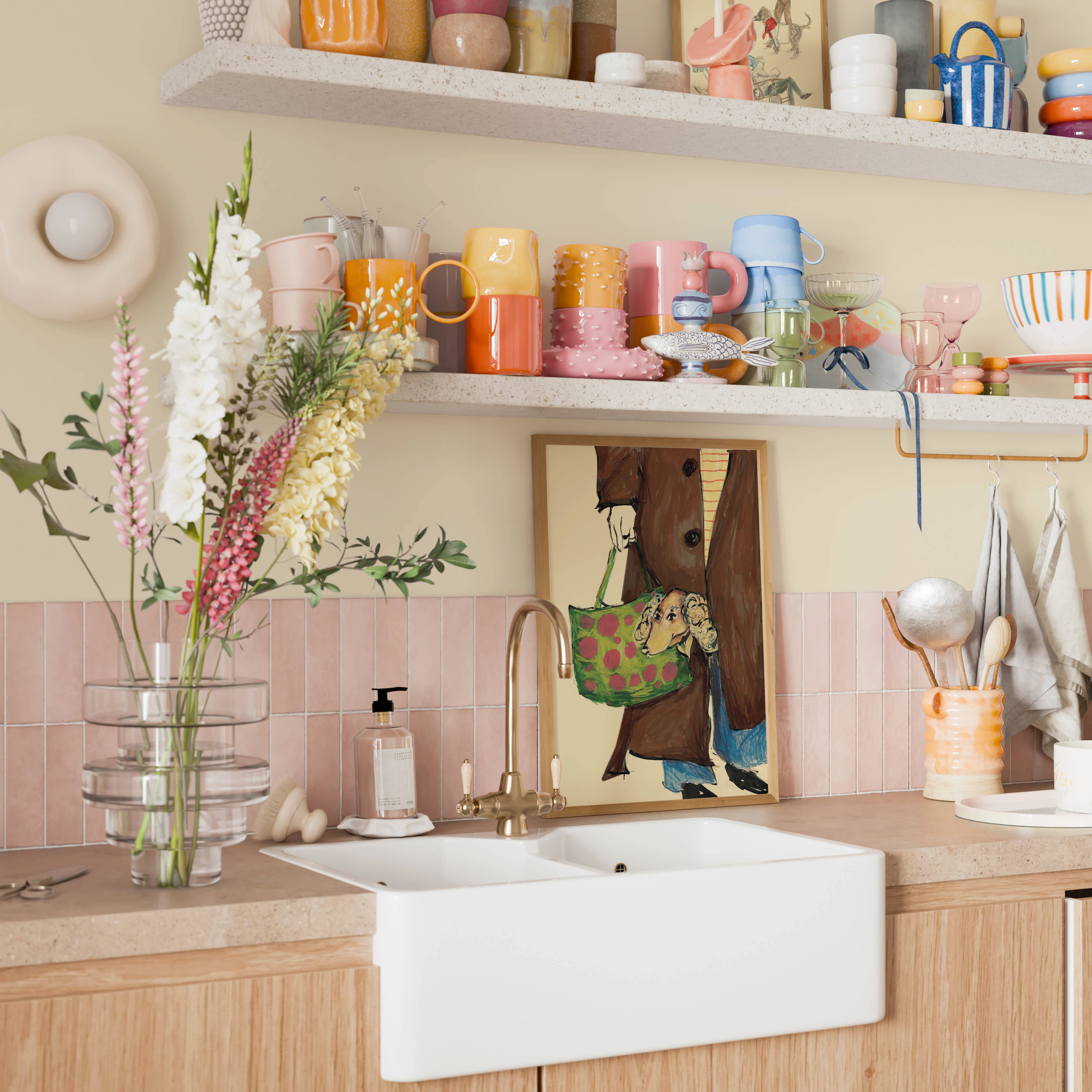 A bright kitchen scene with a white farmhouse-style double sink, gold faucet, pink tile backsplash, open shelves filled with colorful mugs and dishes, and a framed art print on the wall.