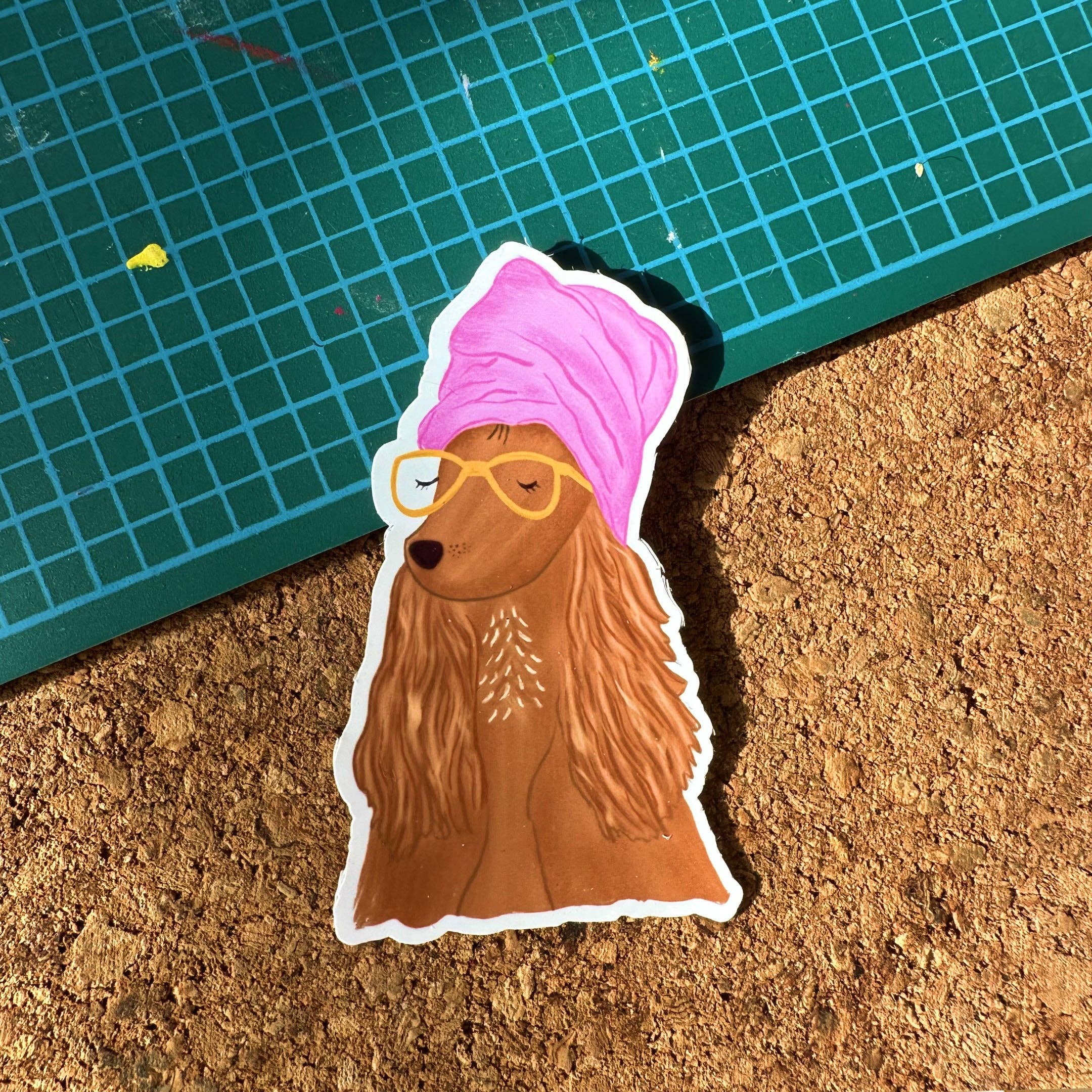 Sticker of a dog with a pink bandana and yellow glasses on a cork surface with a green mat.