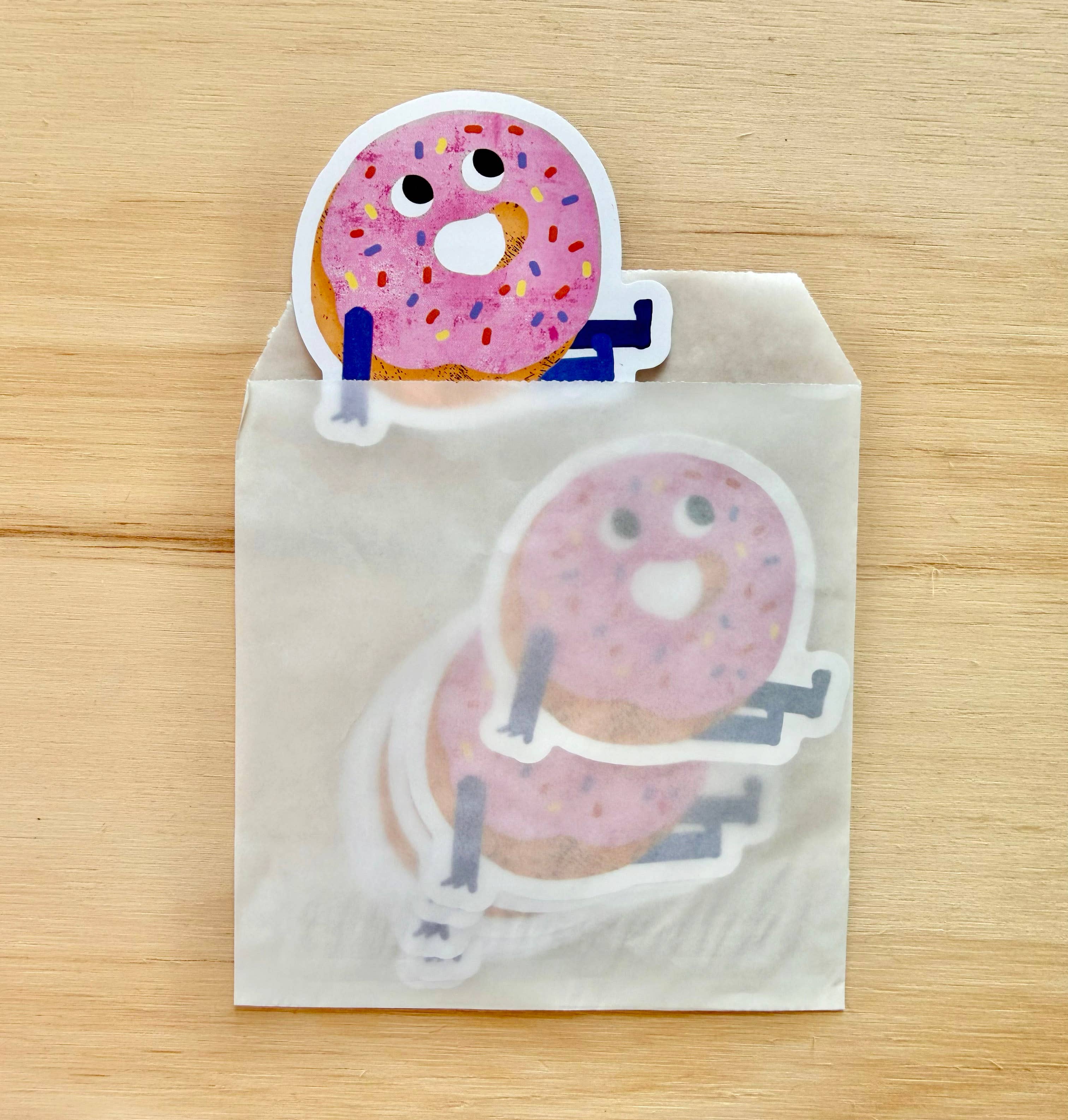 A stack of die-cut cartoon donut stickers with pink frosting and colorful sprinkles, partially sticking out of a clear plastic bag on a light wood surface.