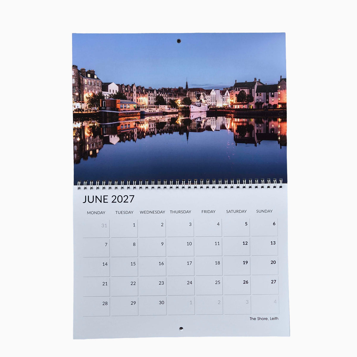 A single page spiral bound desk calendar showing June 2027 with a printed photograph of a waterfront town at dusk and the days of the week below, photographed against a plain white background.