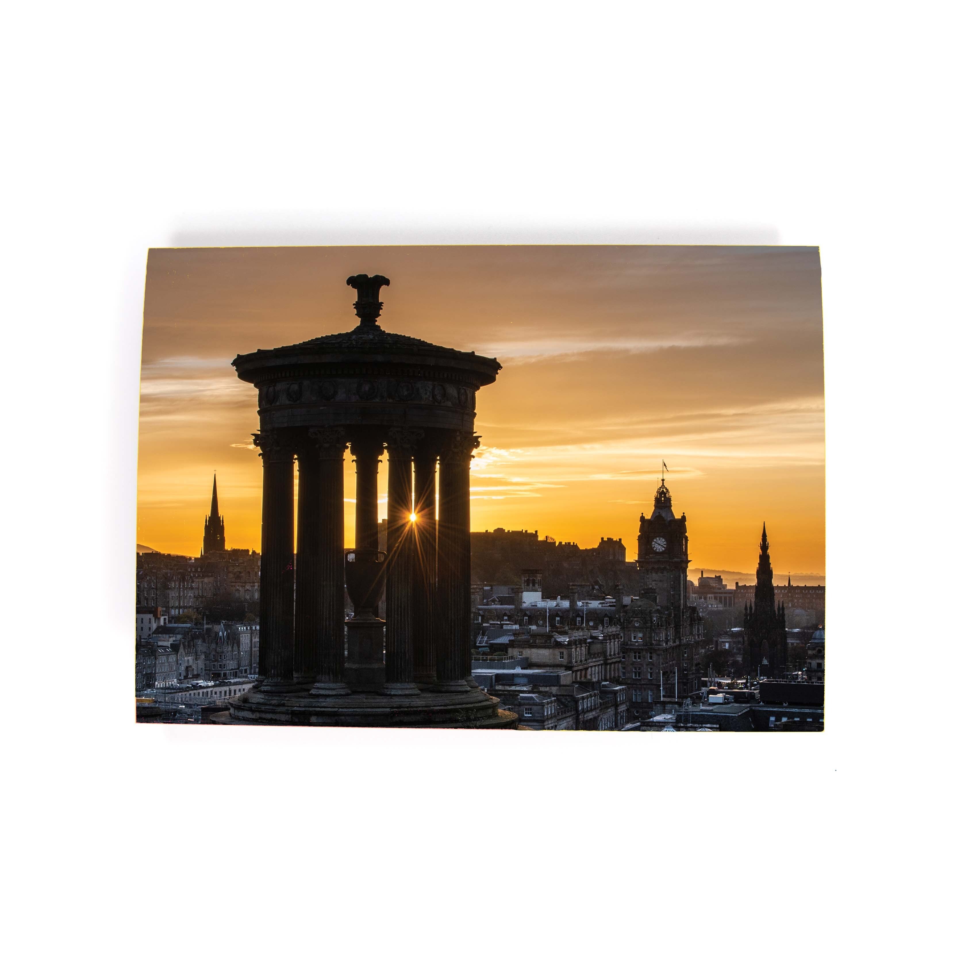 An A6 greeting card featuring a photograph of the Edinburgh skyline from Calton Hill during sunset, with a silhouette of a monument and the sun setting behind it.