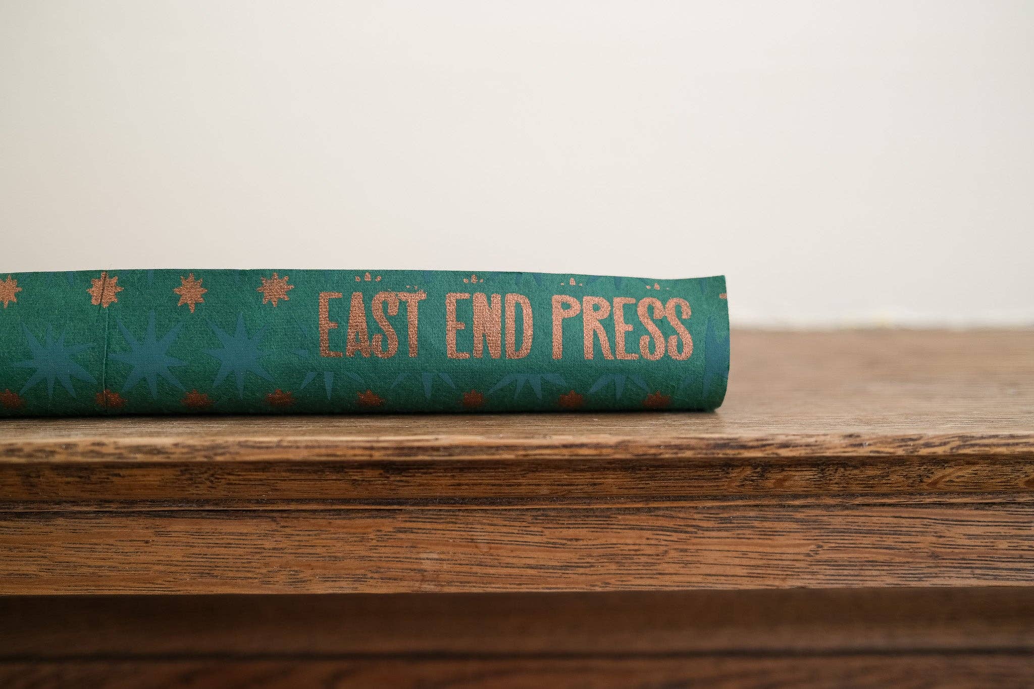 A close-up of a teal wrapping paper with gold text reading 'EAST END PRESS' resting on a wooden surface against a plain light background.