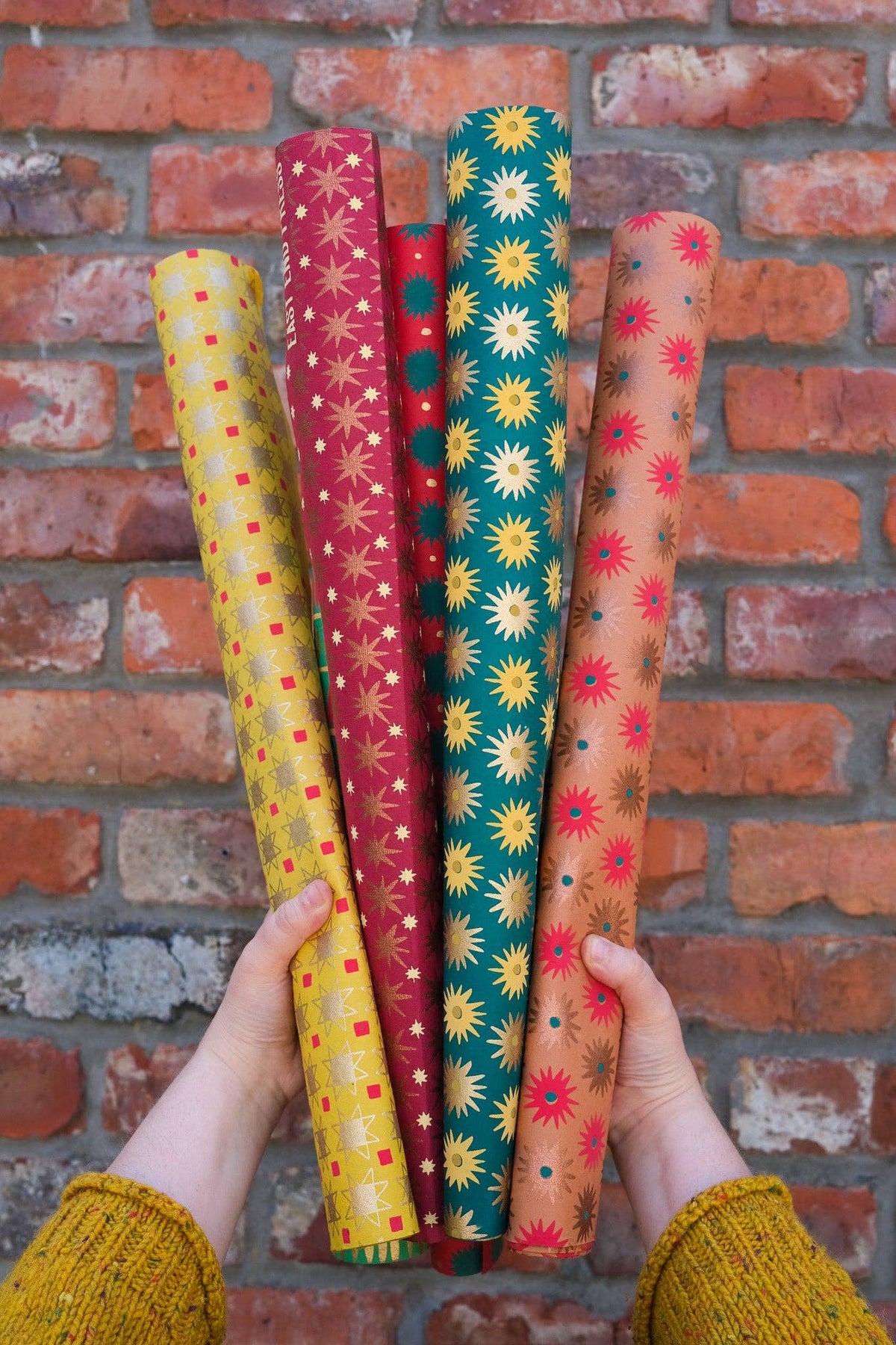 Four rolled sheets of patterned wrapping paper held up by two hands against a red brick wall. The wrapping paper features retro starburst and polka dot designs in yellow, red, teal, and peach tones.