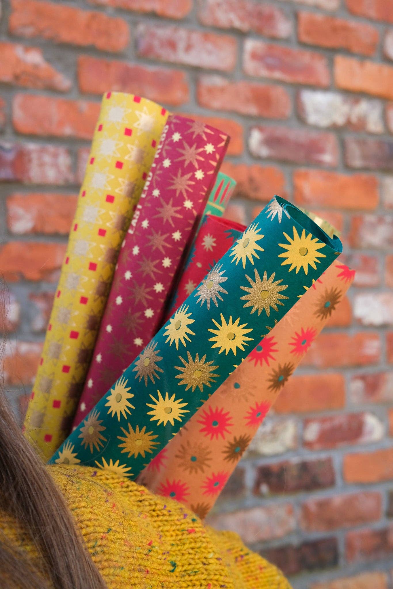 Four rolls of patterned wrapping paper in yellow, red, teal, and orange with starburst and geometric designs, held up against a red brick wall.