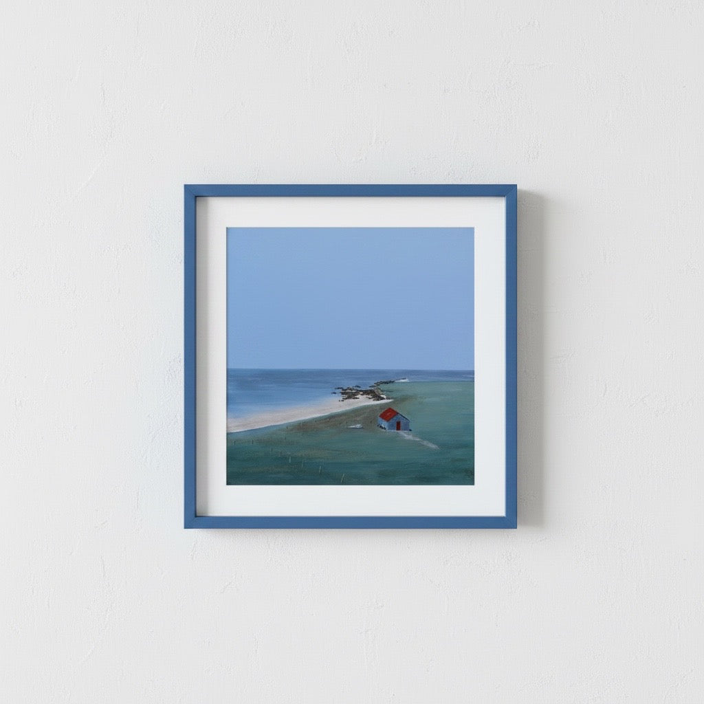 A framed art print depicting a coastal landscape with a small red-roofed house on a sandy spit, mounted on a textured white wall.