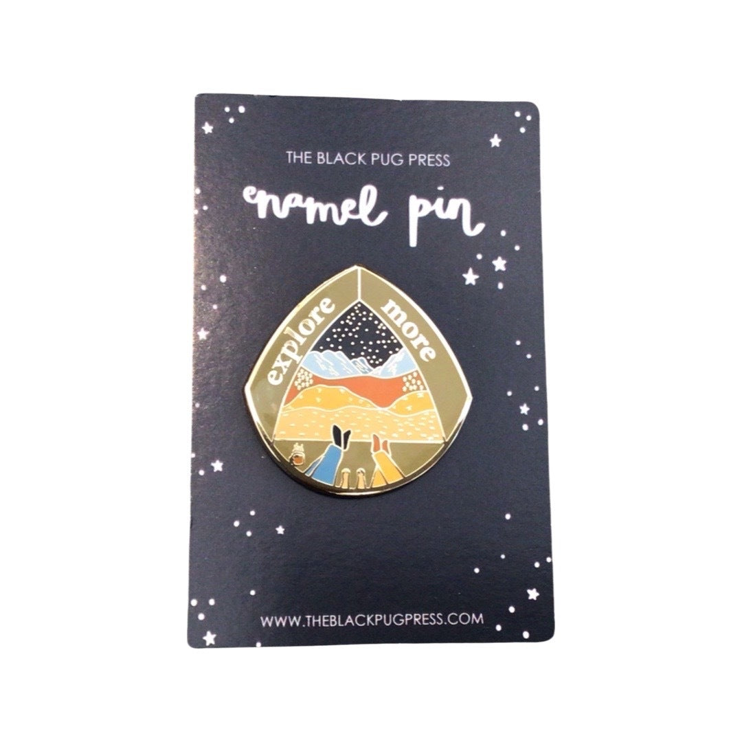 'Explore More' enamel pin badge, featuring an illustration of a couple and their dog looking out from their tent over mountains and getting ready to explore!