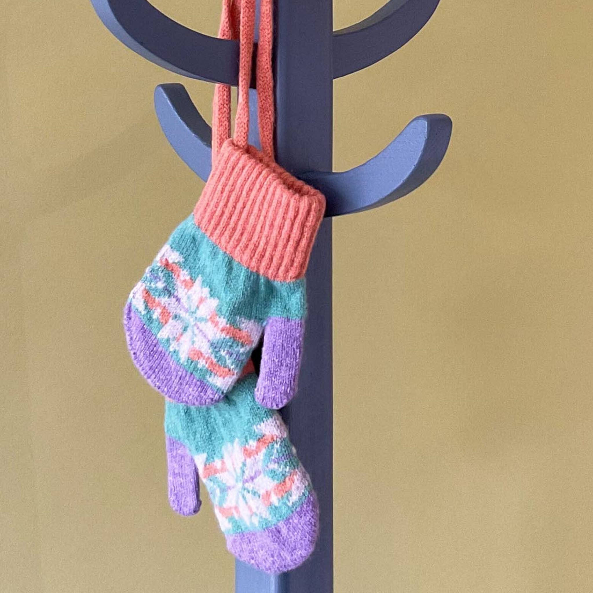 Colourful knitted mittens hanging on a dark coat rack against a beige background.