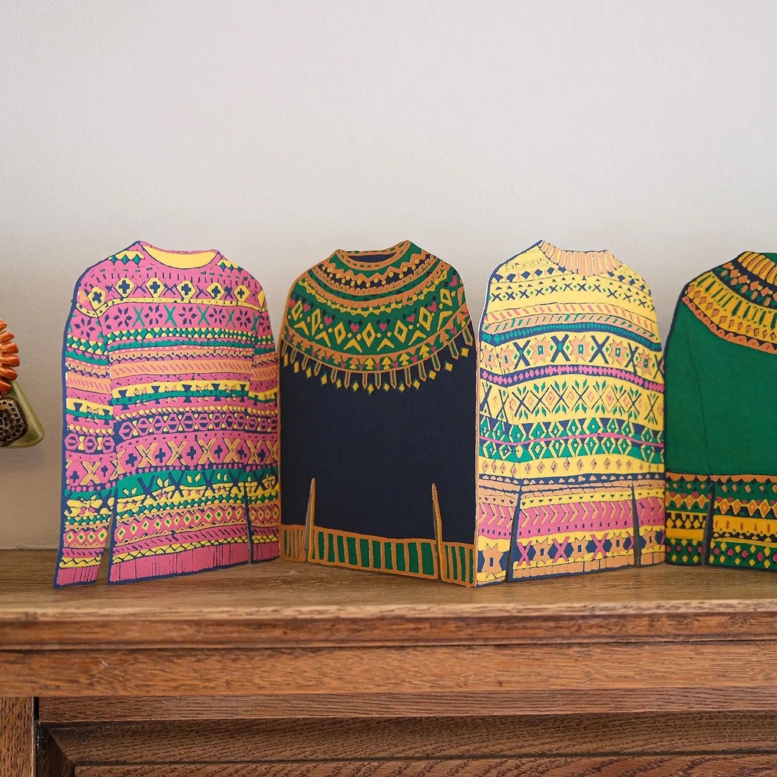 This Fairisle sweater concertina garland features 4 repeated pink, yellow, green and navy patterned sweaters standing on a wooden shelf.