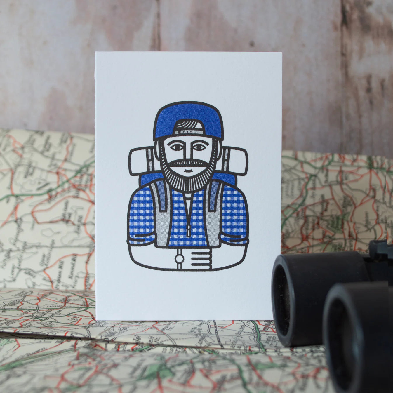 Illustration of a person with a backpack on a white card, sat on a map with binoculars in the foreground.
