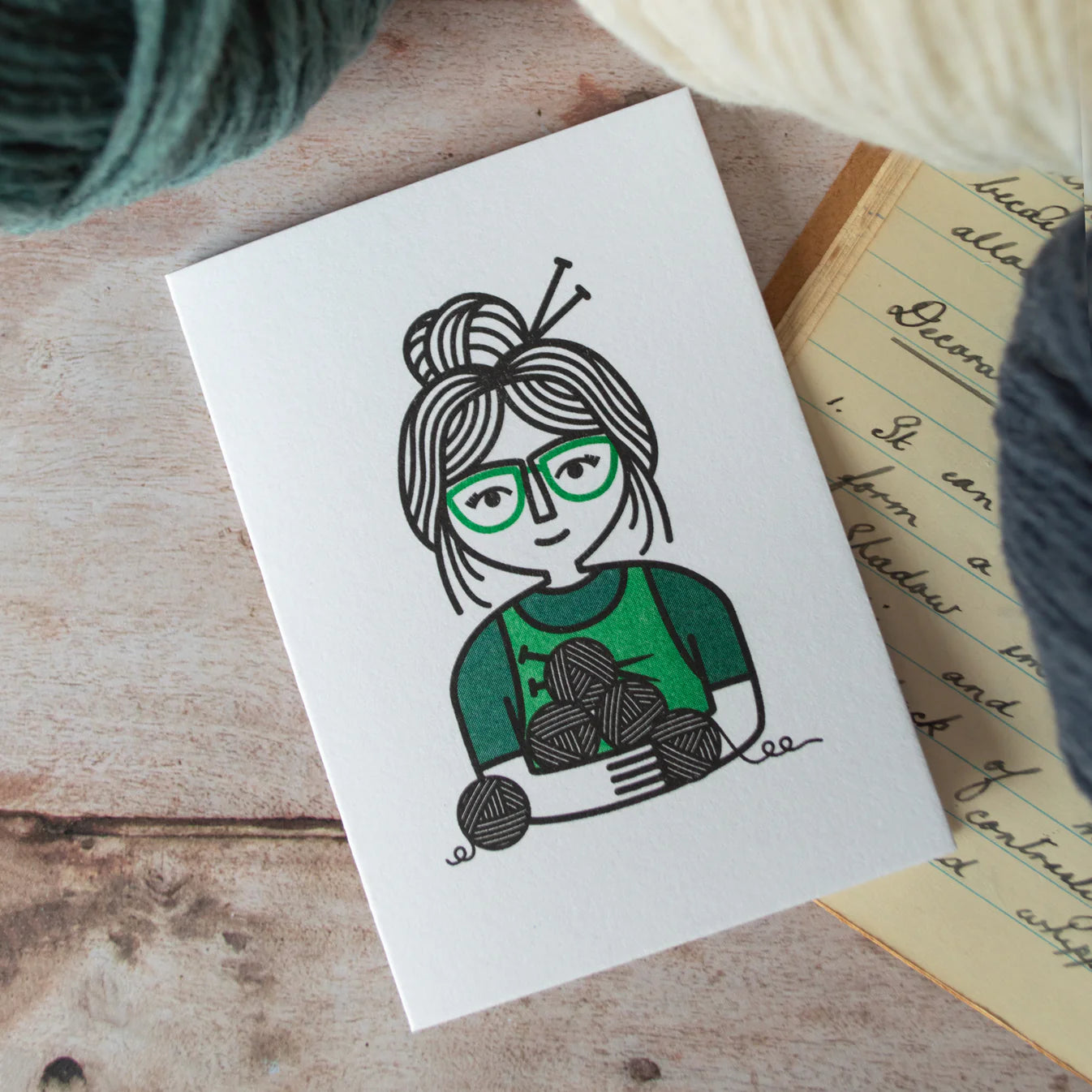 Card with a illustration of a person wearing glasses and holding yarn on a wooden surface, with yarn scattered around.