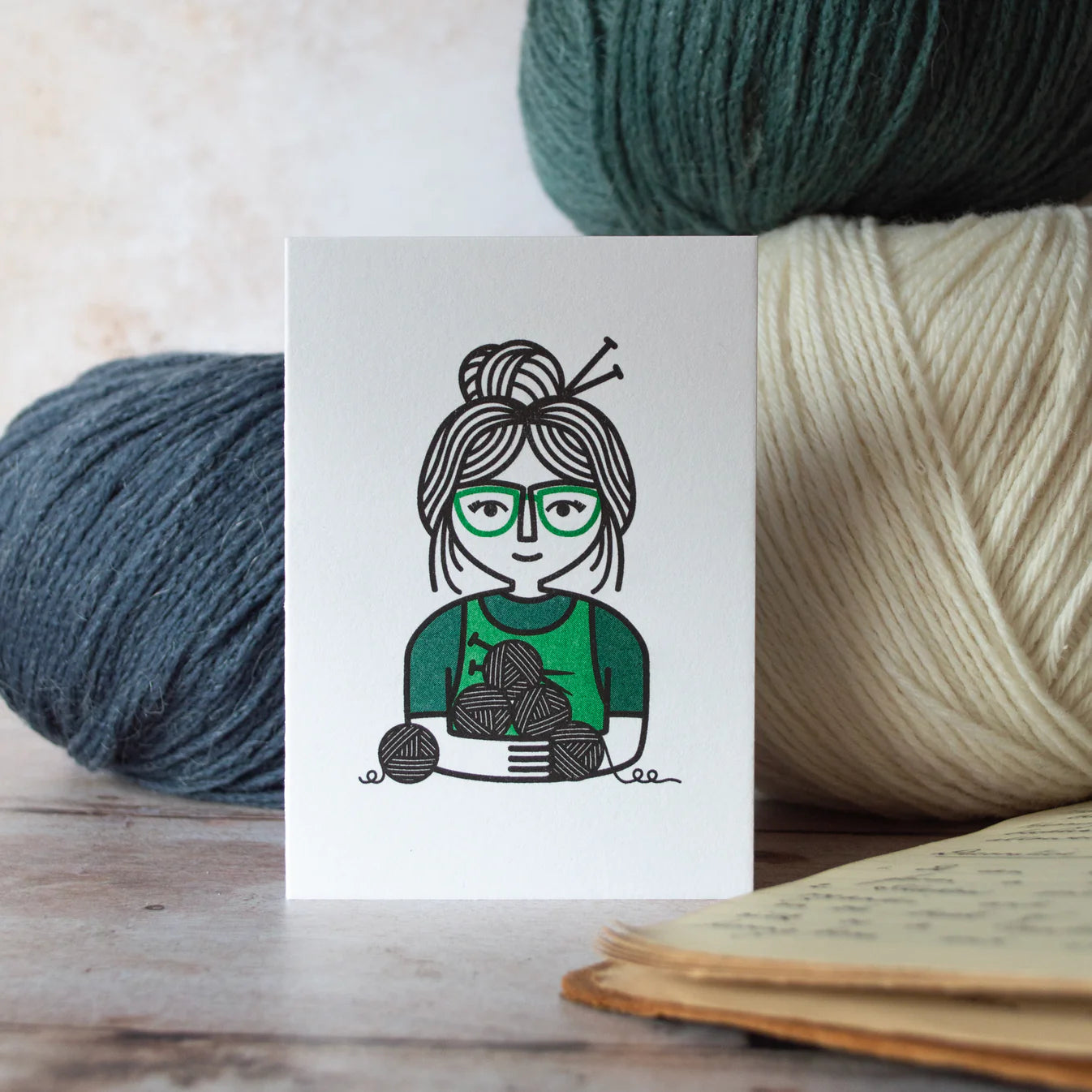Card with a illustration of a person holding yarn and surrounded by yarn balls on a wooden surface.