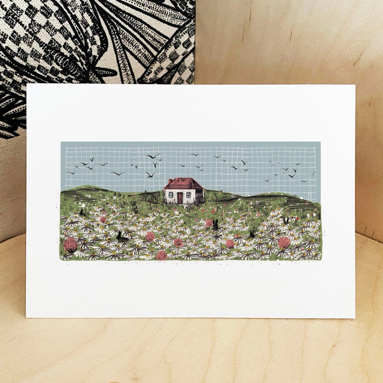 Greeting card with a house in a field of flowers and birds on a checkered background.