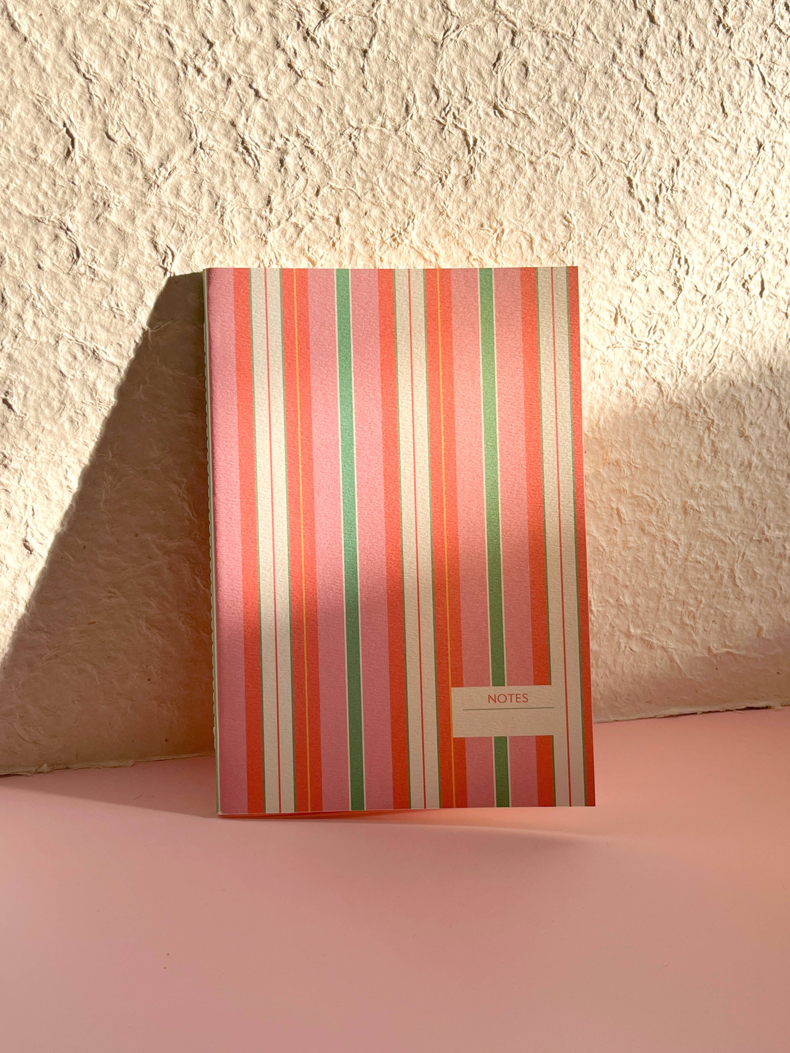 A notebook with vertical pink, orange, green, and white stripes, standing on a pink surface against a textured beige wall.