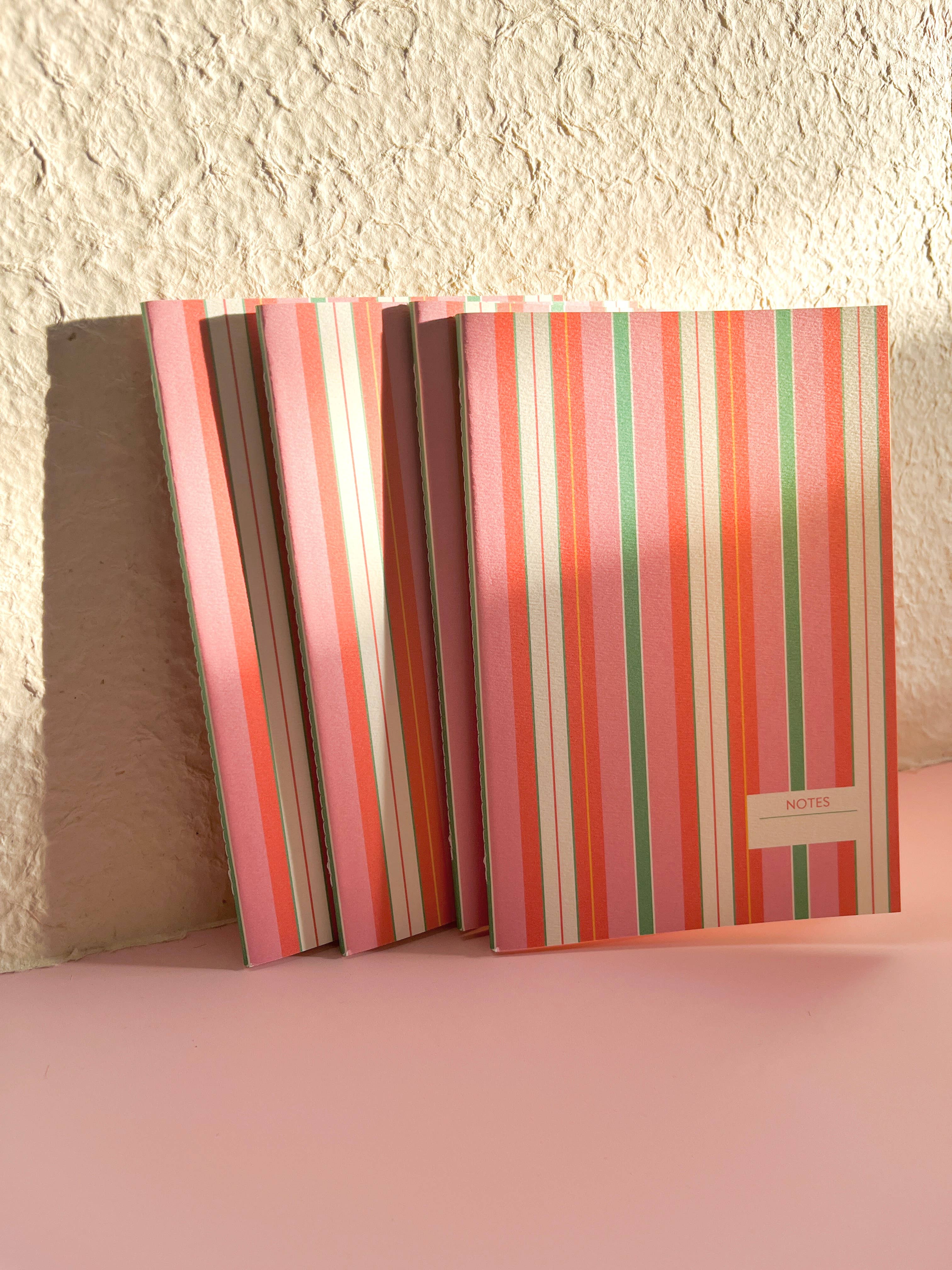 A stack of striped notebooks or journals with pink, coral, peach, green, and cream vertical stripes, leaning against a textured beige wall on a pink surface.