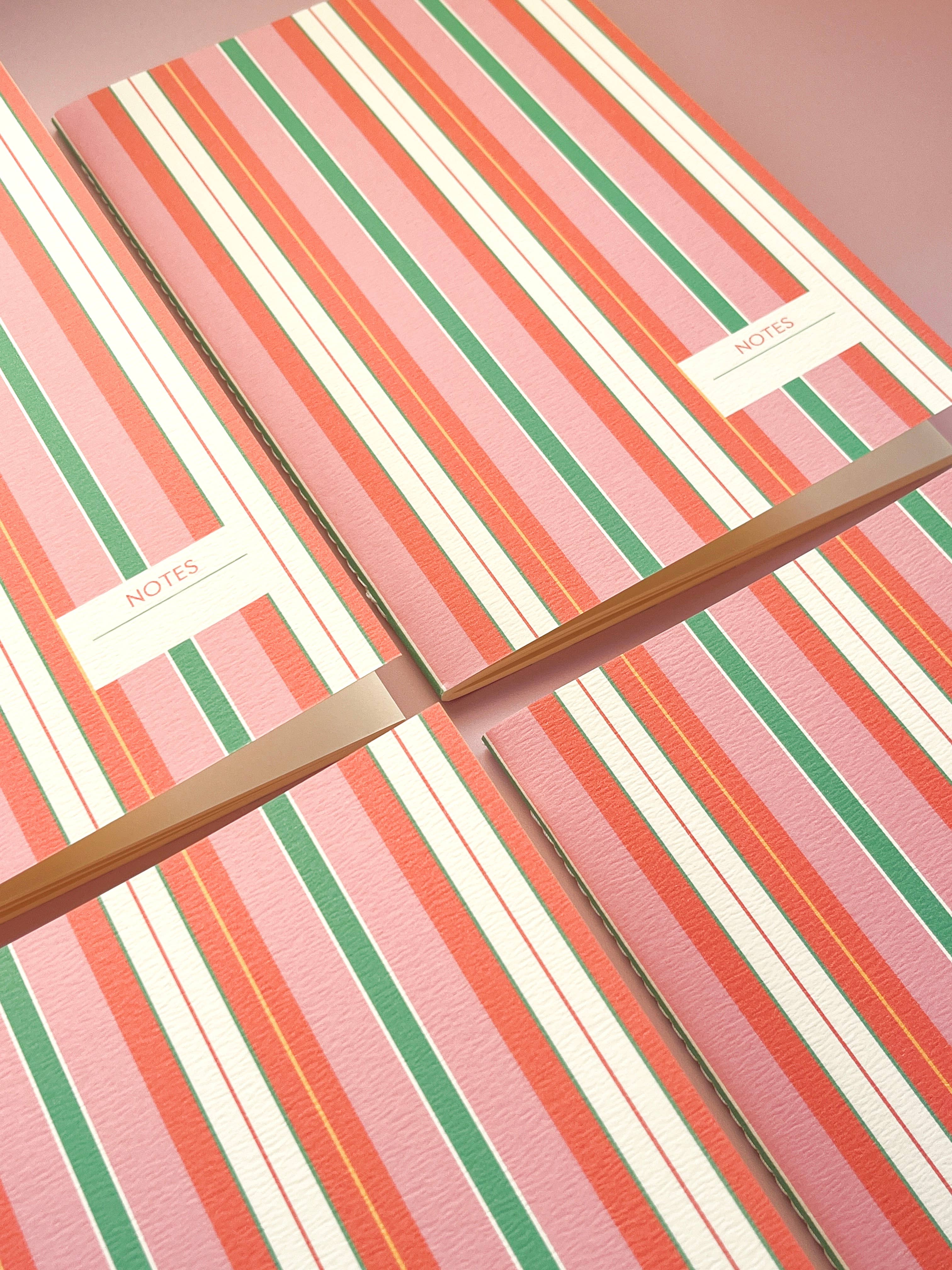 Four striped hardcover notebooks with pink, orange, green, and white vertical stripes, arranged diagonally on a matching pink background.
