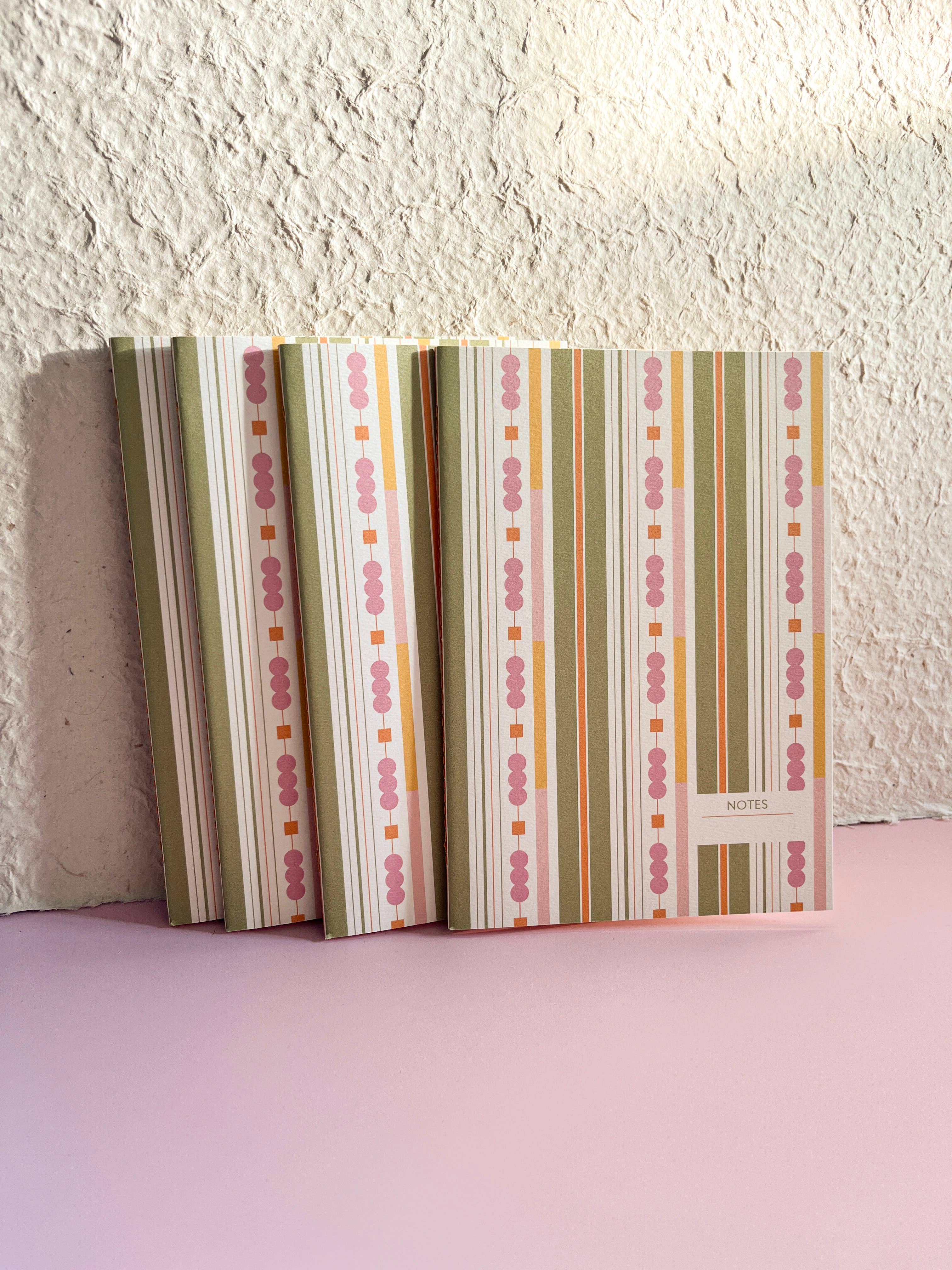 A set of patterned notebooks or journals with vertical stripes in sage green, pink, orange, and cream, standing upright against a textured off-white wall on a pink surface.