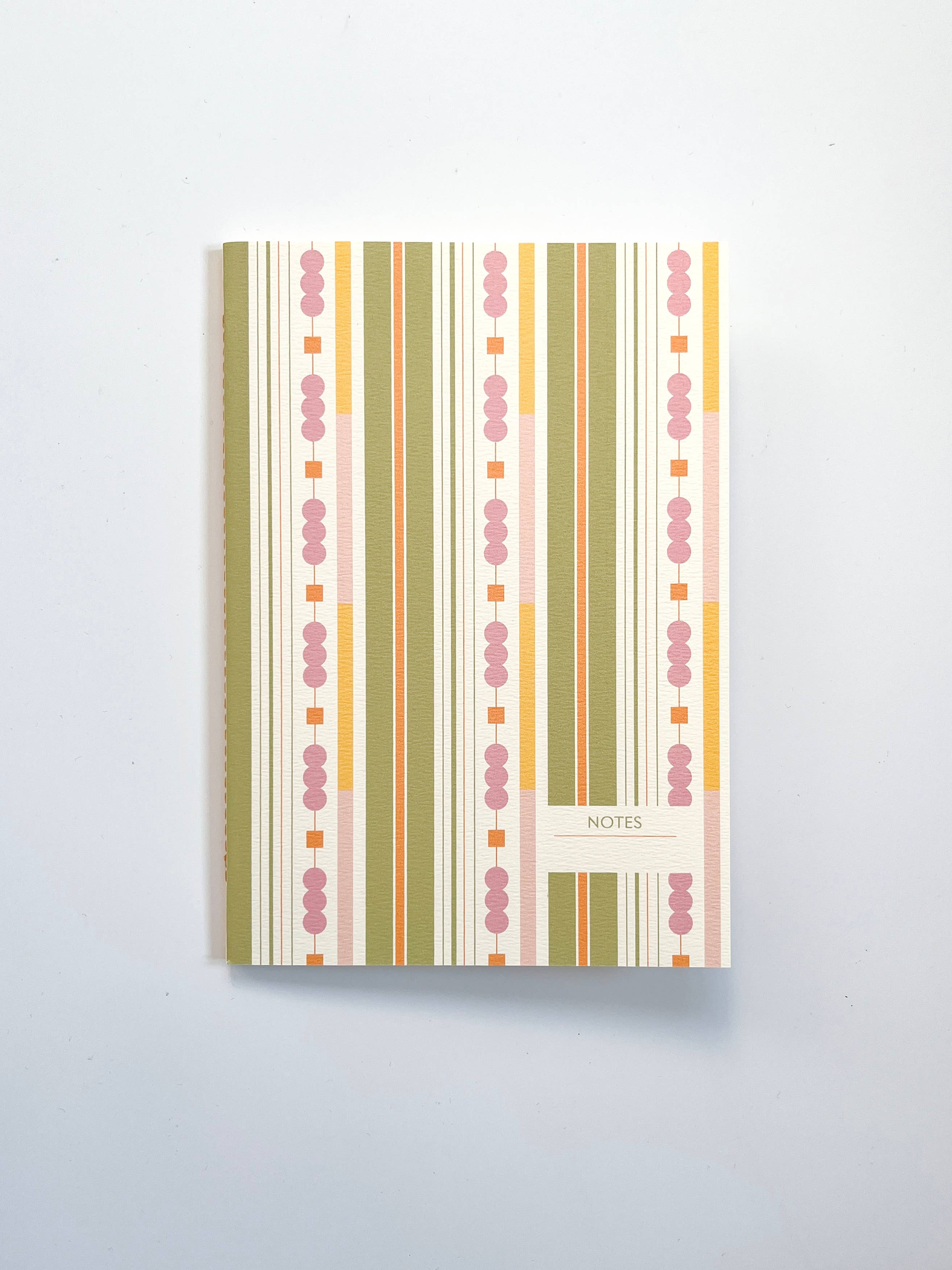 A vertical notebook or journal with a colourful striped cover pattern in pink, yellow, green, and orange on a cream background, featuring a small label that reads 'NOTES' at the bottom right.