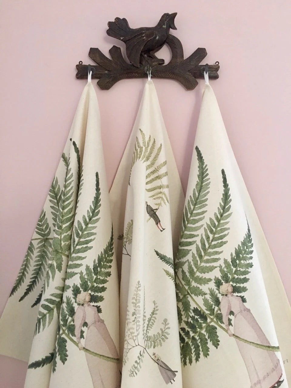 Set of three decorative tea towels with nature-themed designs hanging on decorative hooks against a pink wall.