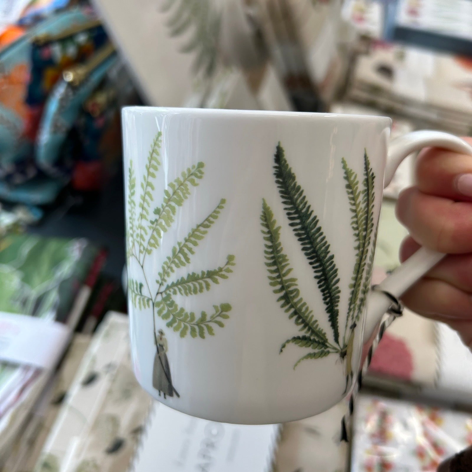 White mug with green leaf patterns held by a hand in a store setting.