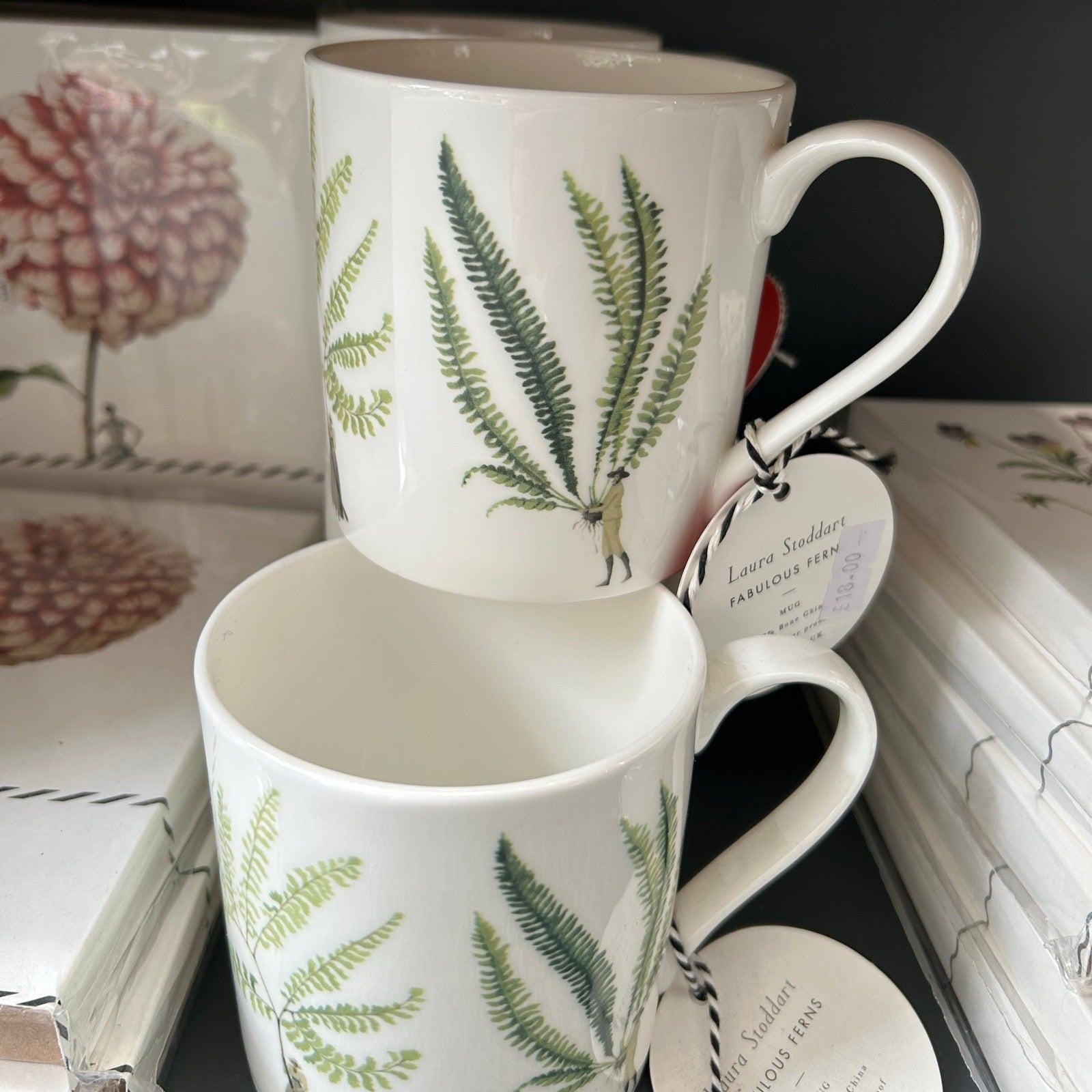 Two white mugs with green leaf patterns on a surface with notebooks in the background.