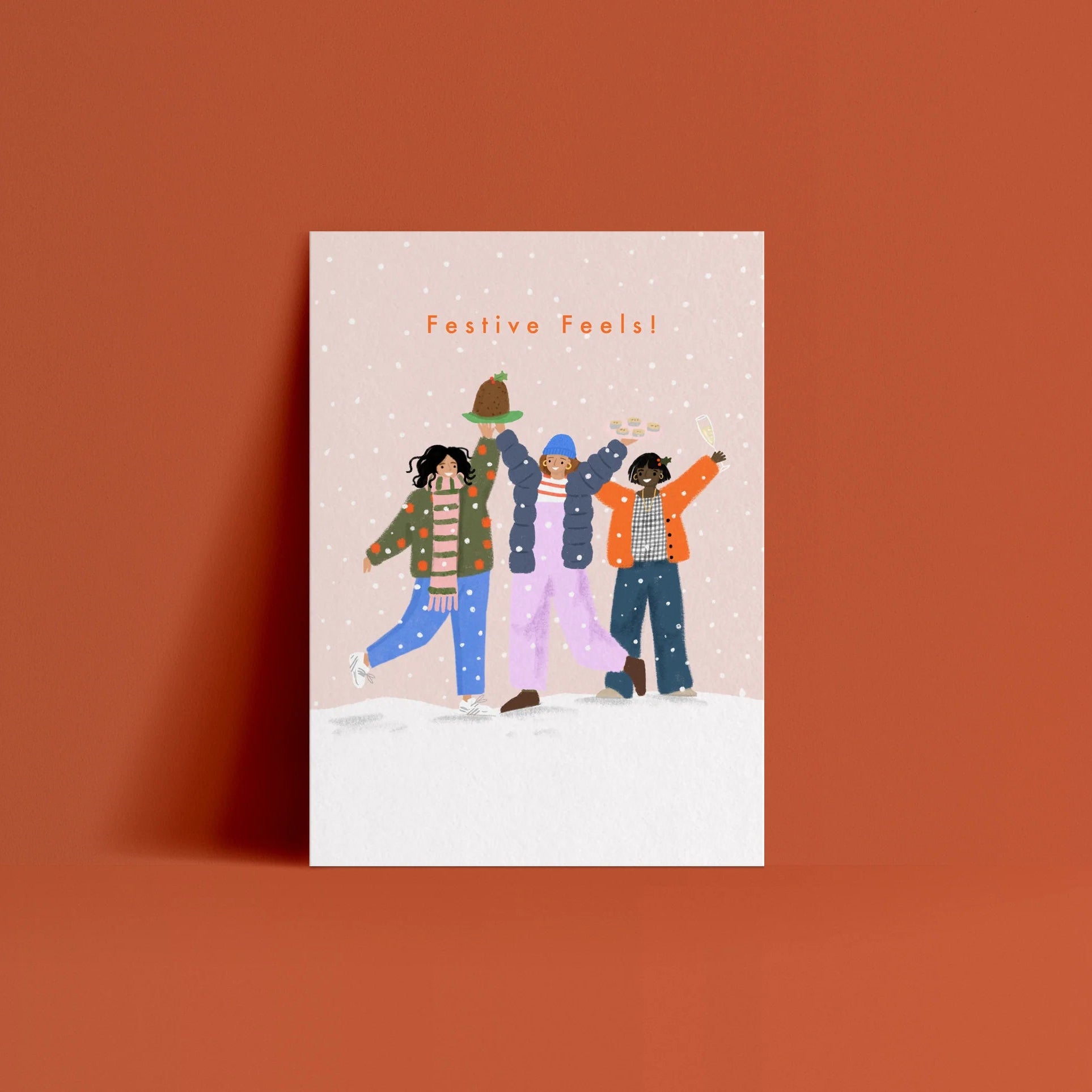 A vertical greeting card featuring a festive illustration of three women in winter clothing under the text 'Festive Feels!', displayed against a solid terracotta orange background.