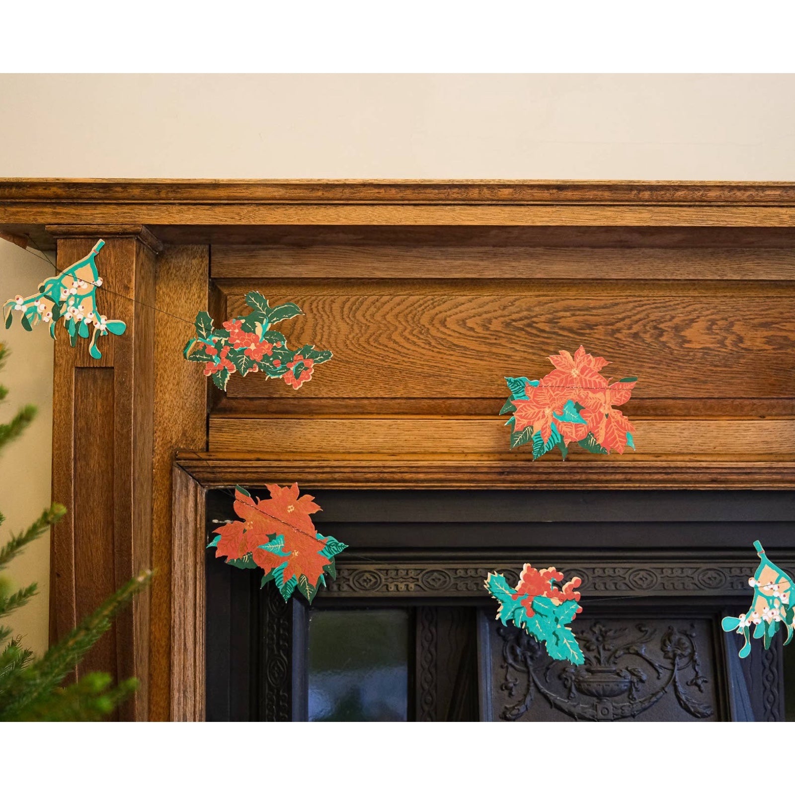 Festive plant garland features ten screen-printed plants including holly, red berries, mistletoe, ivy and poinsettias in shades of green, red and gold hanging on a wooden fireplace.