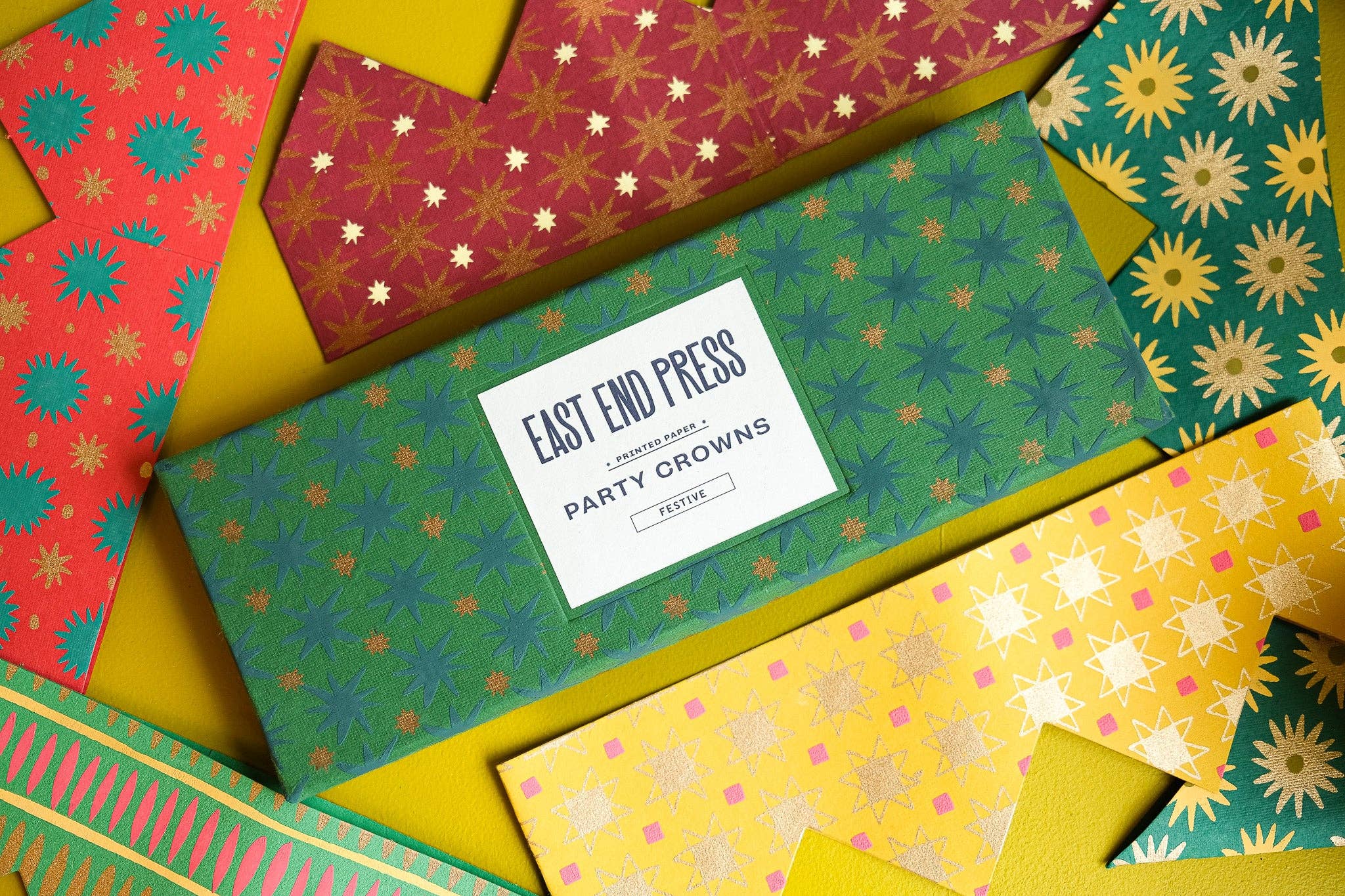 A flat-lay of multiple colorful patterned party crowns by Easy End Press, arranged on a bright yellow background. The central crown is dark green with a starburst pattern and a white label reading 'EAST END PRESS PARTY CROWNS'.