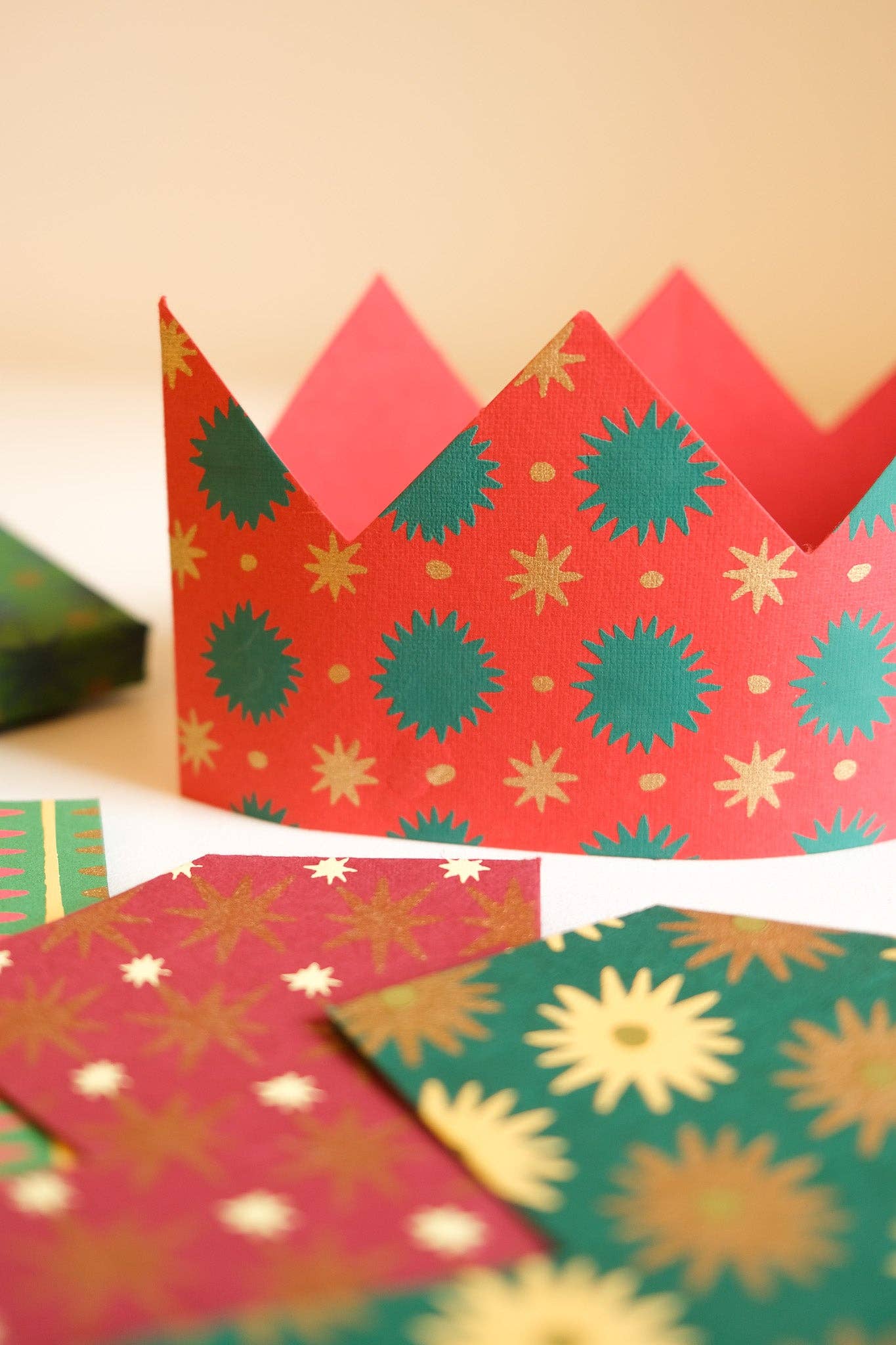 A festive paper crown with red, green, and gold holiday patterns, shown alongside matching patterned paper crowns on a light beige background.