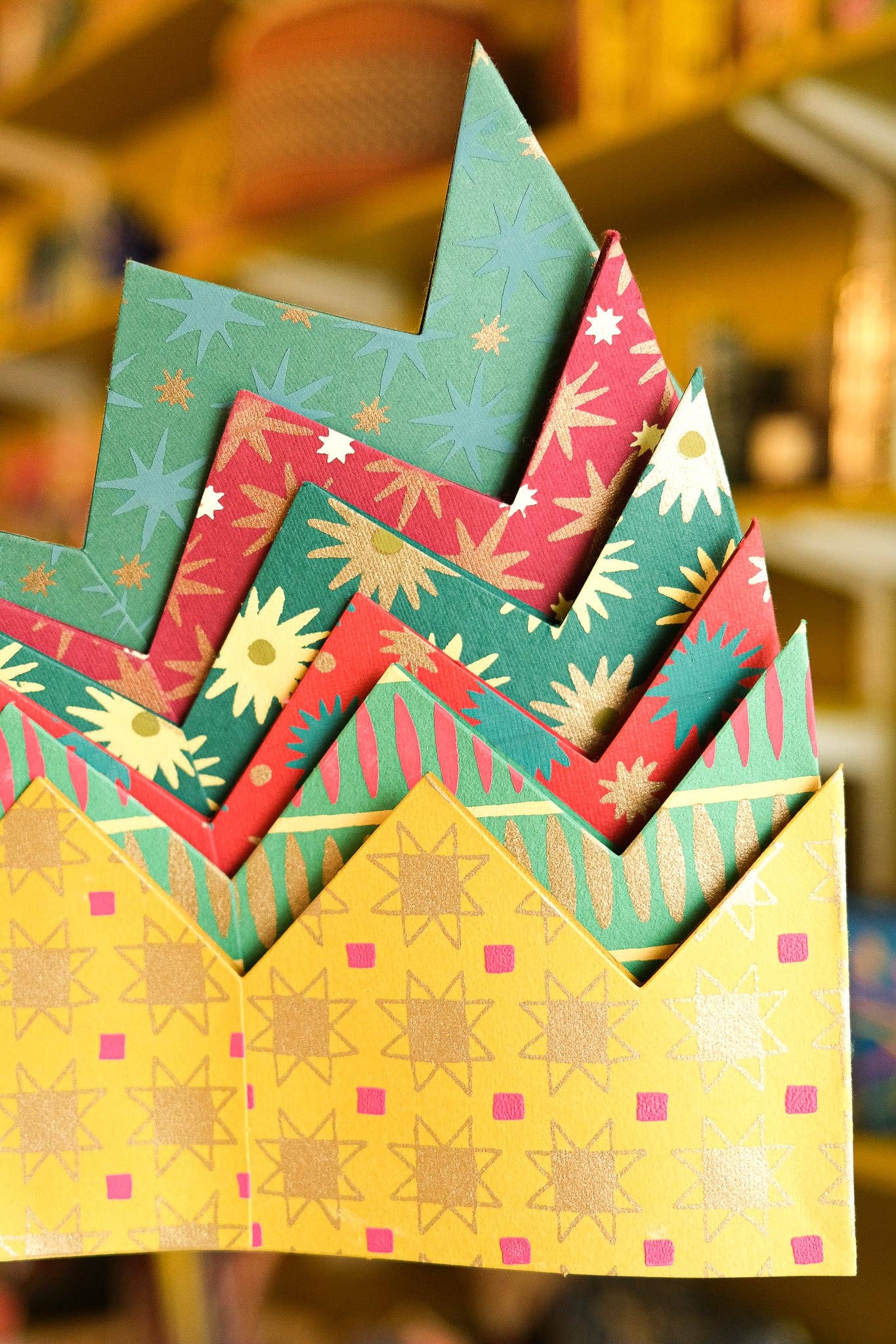 A close-up of a stack of colorful patterned paper crown hats in yellow, green, and red, each with geometric star and flower designs, set against a warm, blurred background.