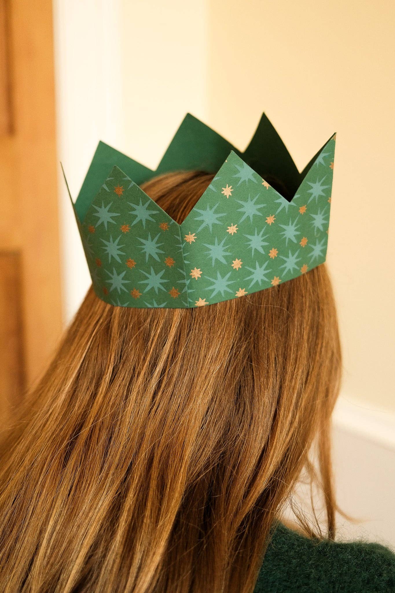 A green paper crown with a repeating snowflake pattern, worn on the back of a person's head with long light brown hair.