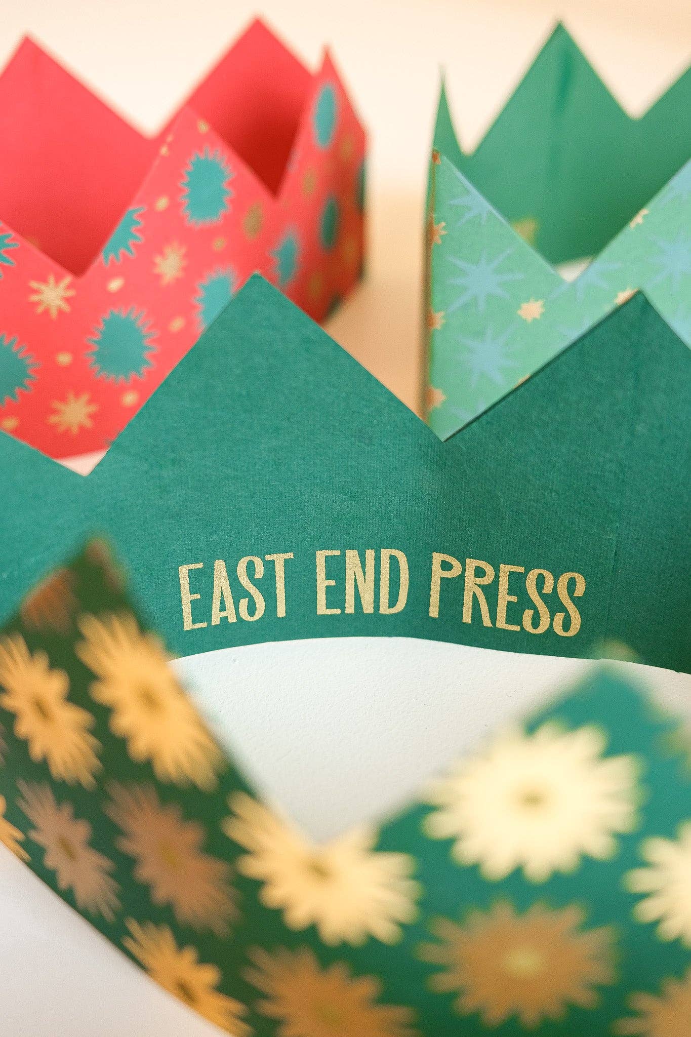 Three paper crown party hats with pointed peaks, shown close up on a light background. The front crown is dark green with gold foil text reading 'EAST END PRESS' and a gold floral pattern along the lower edge.
