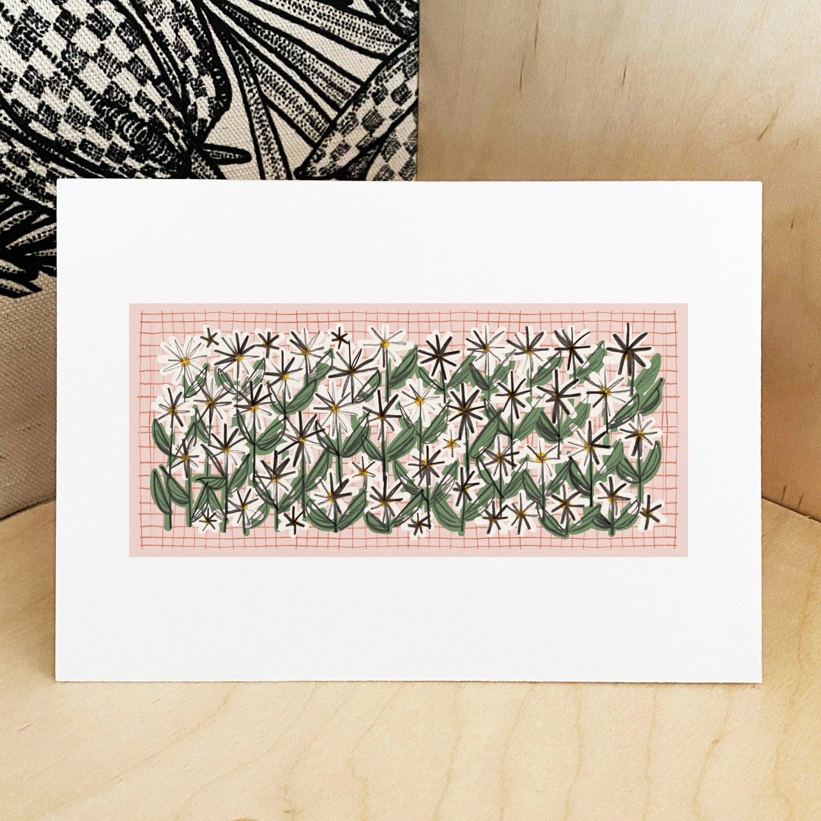 Art print with floral design on a pink grid, displayed on a wooden surface.