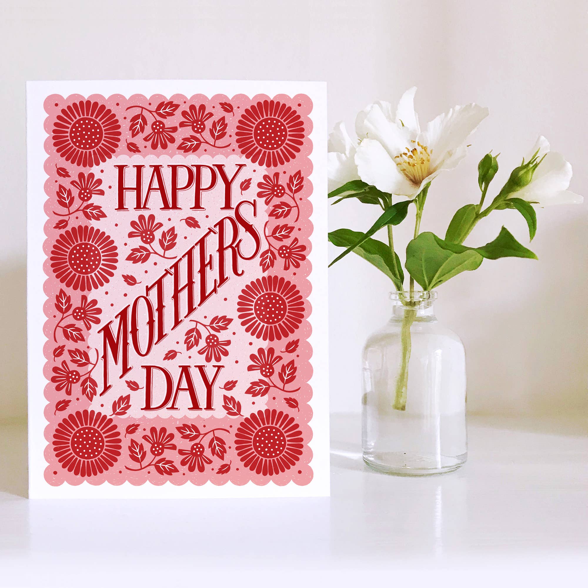 Card with 'Happy Mother's Day' text and floral design next to a vase with white flowers on a white background.