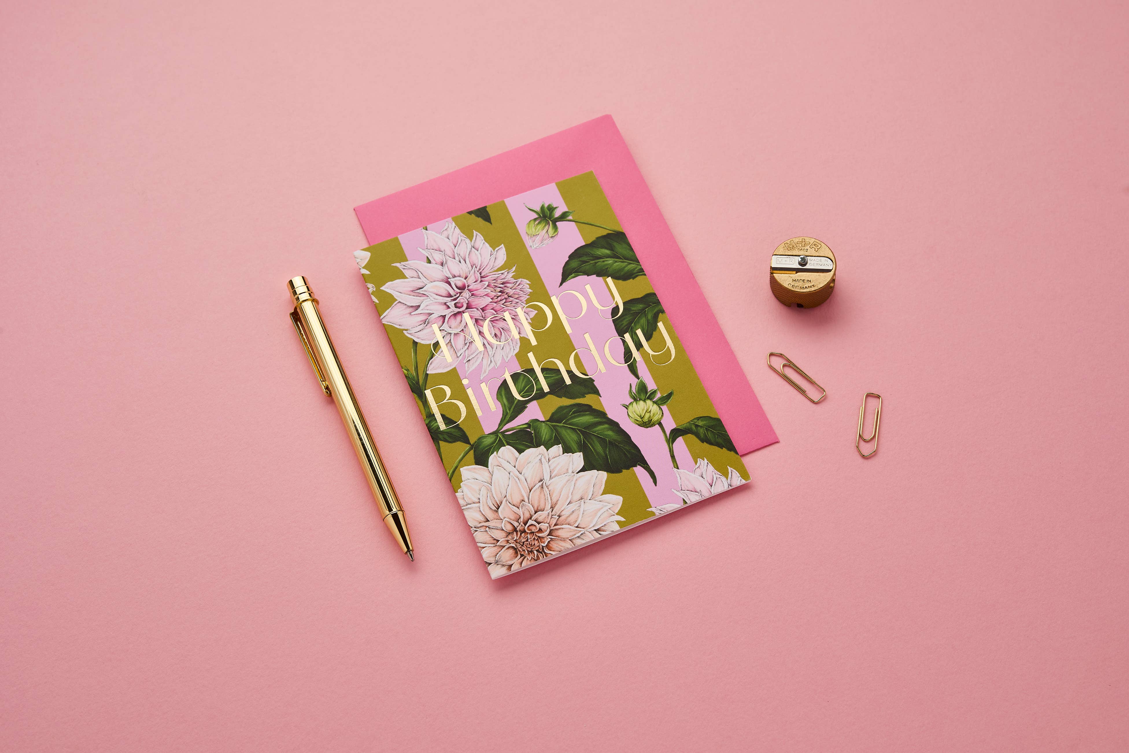 A floral 'Happy Birthday' greeting card with a pink envelope, styled in a flat lay on a pink background with a gold pen, paper clips, and a small gold box.